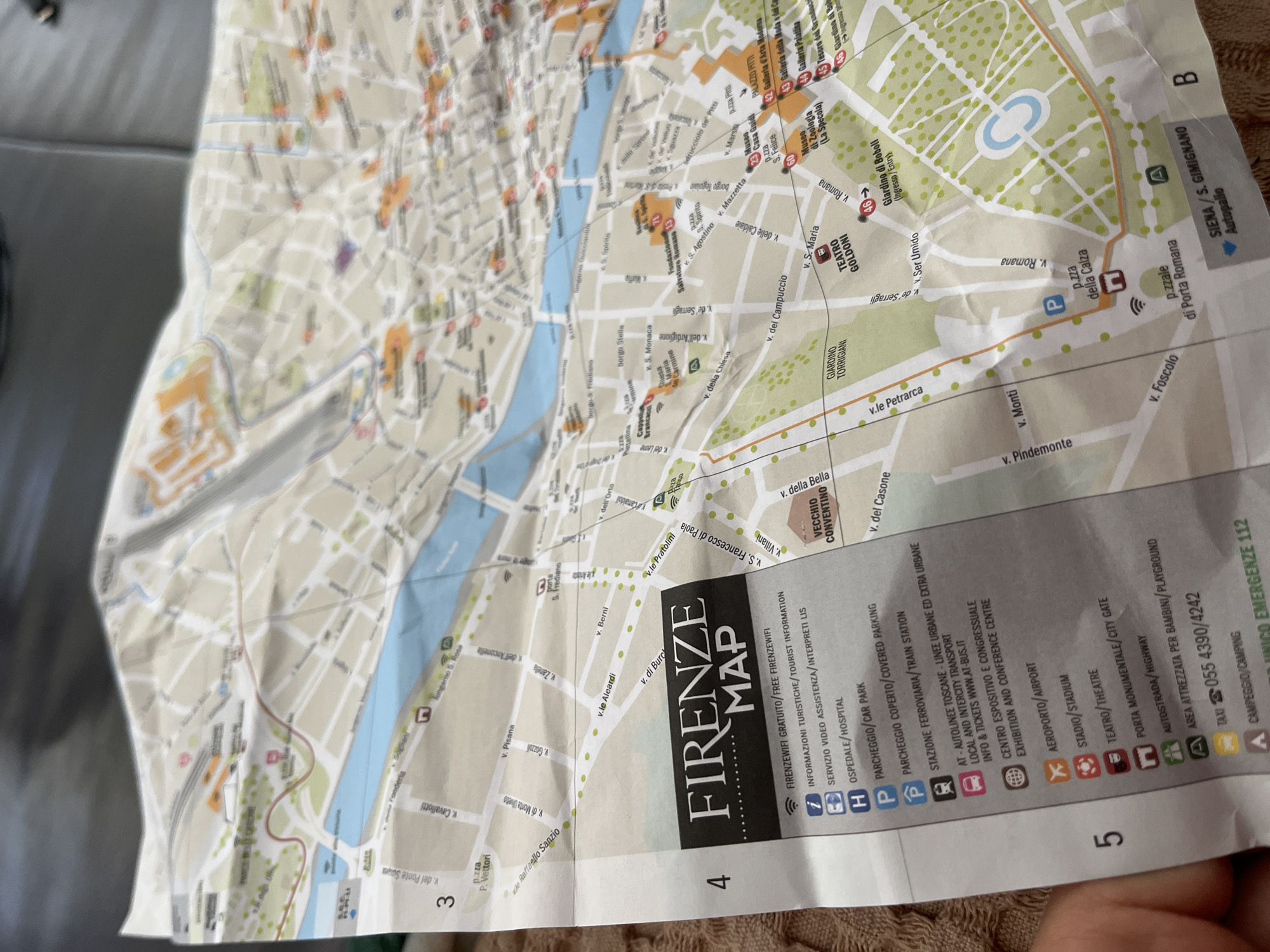 The image captures a crumpled paper map of Florence, Italy, held by a person whose hand is partially visible in the bottom right corner. The map, titled "FIRENZE MAP," is oriented diagonally, indicating it is actively being consulted. Its creased and worn condition suggests frequent use.
The map displays detailed street layouts, green areas representing parks such as "Giardino di Boboli" and "Giardino Torrigiani," and the blue ribbon of the Arno river. Various landmarks and points of interest are marked with icons and text. Notable locations include "PALAZZO PITTI," "TEATRO GOLDONI," "Museo di Zoologia (La Specola)," and "Galleria d'Arte Moderna." Street names like "v. Romana," "v.le Petrarca," and "v. del Campuccio" are also visible.
A comprehensive legend on the right side lists symbols for essential services and attractions, including "FIRENZEWIFI GRATUITO / FREE FIRENZEWIFI," "INFORMAZIONI TURISTICHE / TOURIST INFORMATION," "OSPEDALE / HOSPITAL," and various parking options. Transportation details cover "STAZIONE FERROVIARIA / TRAIN STATION," local and intercity transport ("AT - AUTOLINEE TOSCANE"), and "AEROPORTO / AIRPORT." Other entries include "CENTRO ESPOSITIVO E CONGRESSUALE," "STADIO / STADIUM," "TEATRO / THEATRE," "PORTA MONUMENTALE / CITY GATE," "AUTOSTRADA / HIGHWAY," "AREA ATTREZZATA PER BAMBINI / PLAYGROUND," "TAXI 055 4390/4242," "CAMPEGGIO / CAMPING," and "UNICO EMERGENZE 112." The map's grid coordinates, "3," "4," "5," and "B," are visible along the edges.
While the map explicitly details the city of Florence, Italy, the image itself was captured in Zlatograd, Bulgaria, as per the provided context.