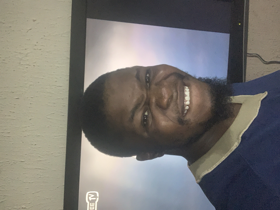 A man is smiling at the camera while on television. He has short black hair, a dark beard, and brown eyes. He is wearing a blue shirt with a tan collar. Behind him is a television set with a cloudy sky background. The television is mounted on a textured wall. A power cable hangs to the right of the television.