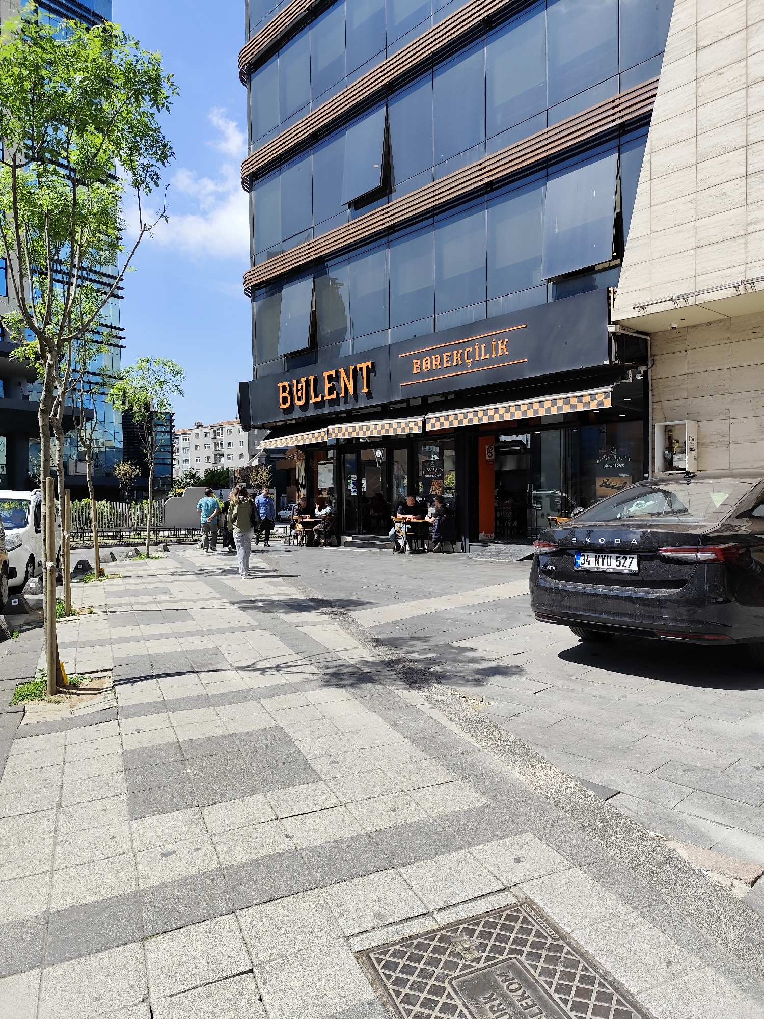 The image shows a street scene in an urban environment. The foreground is dominated by a tiled sidewalk with a sewer grate. A black car is parked on the street, partially blocking the view of a cafe or restaurant named "Bulent Borekcilik". People are seated at tables outside the establishment, while others are walking on the sidewalk. A multi-story building with glass windows and a brown accent borders the restaurant. Trees are present along the sidewalk, adding a touch of greenery to the scene. In the background, other buildings can be seen, indicating a densely populated area. The sky is visible with some clouds. The license plate of the car is 34 NYU 527.