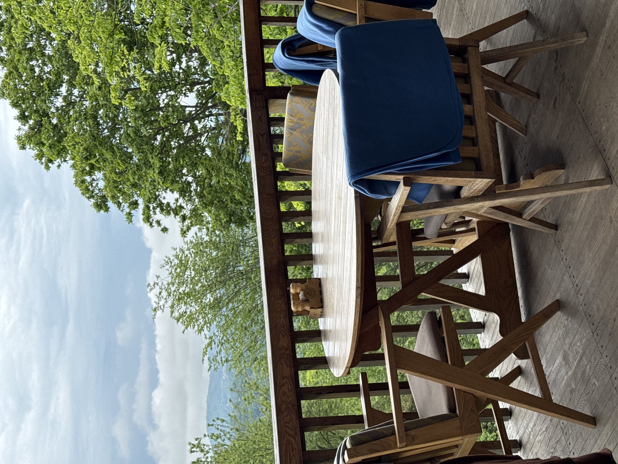 The image shows an outdoor dining area on a porch or balcony with a wooden table and chairs. Blue blankets are draped over some of the chairs. A condiment set rests on the table. The background features a green landscape with trees and a view of mountains under a partly cloudy sky. The location of the image in the city of Progress, Russia is not supported by the visual content.