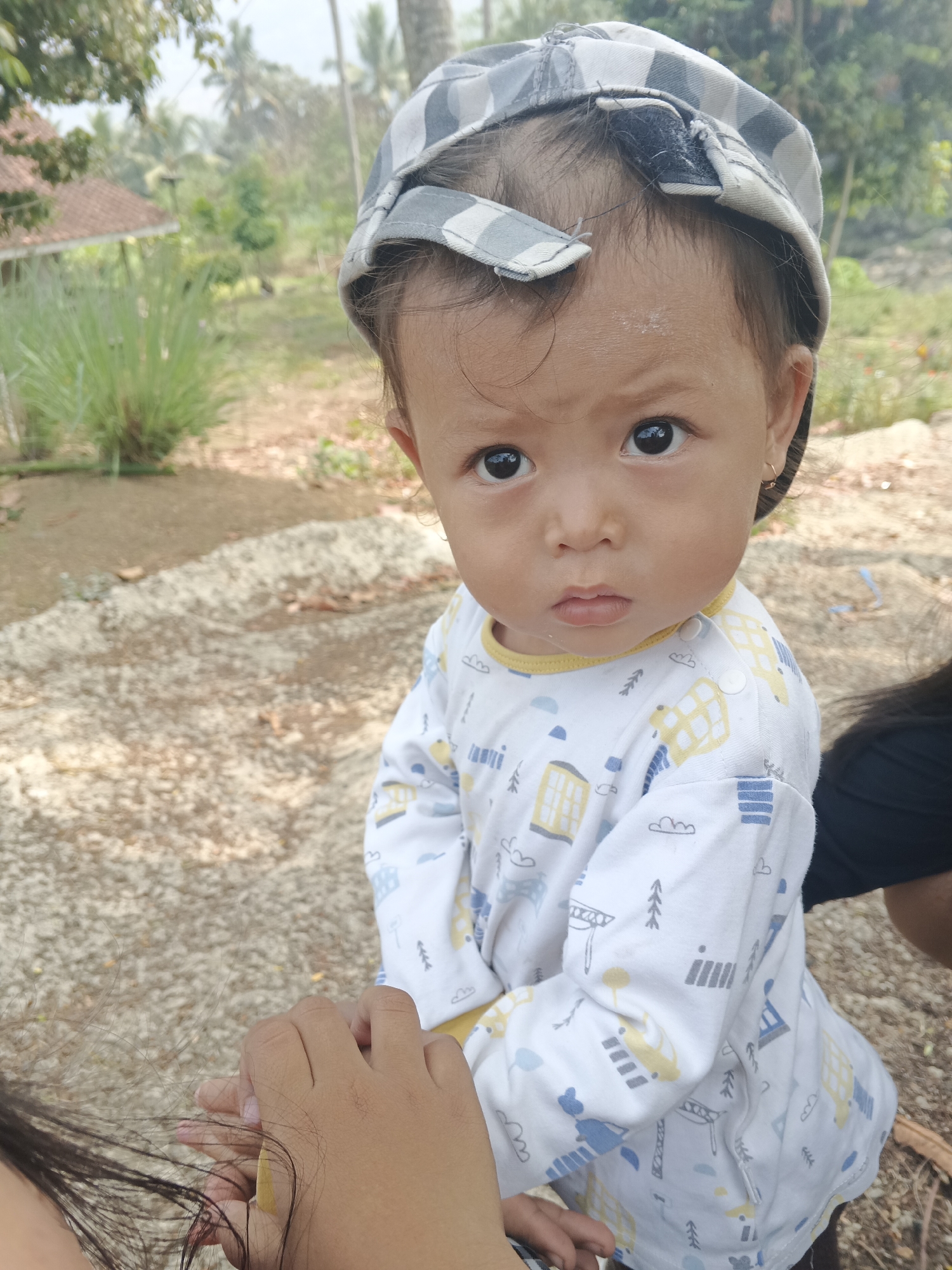 A close-up, eye-level shot captures a young child wearing a grey and white checkered baseball cap and a long-sleeved white shirt with a yellow collar. The shirt is adorned with a playful pattern of yellow cars, grey buildings, and white clouds and trees. The child's dark eyes gaze directly at the camera with a serious expression, and their lips are slightly parted. The child is wearing a small gold hoop earring in their left ear. In the foreground, another person's hands are visible, holding the child's left hand. The background suggests an outdoor setting, possibly in Sukabumi, Indonesia, with a dirt path, some dried leaves, and lush green vegetation. A blurred house with a red roof is visible in the distance. The overall lighting is natural and appears to be daylight.