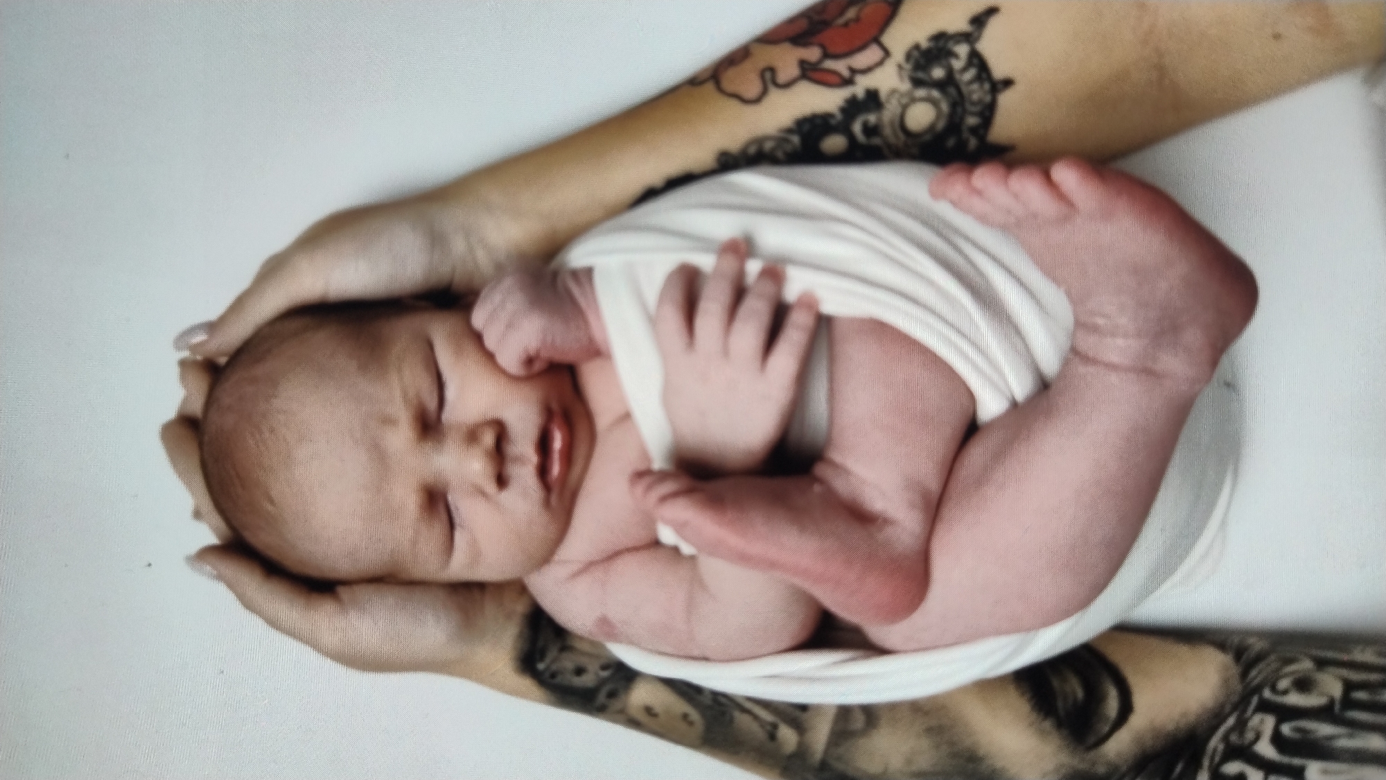 A newborn baby is wrapped in a white cloth and held by two adults. The baby's eyes are closed, and its head is resting on its hands. The adults' arms and hands are visible, as are tattoos on their arms. The background is a plain white wall. There is no discernible location within the city of Mykolaiv, Ukraine.
