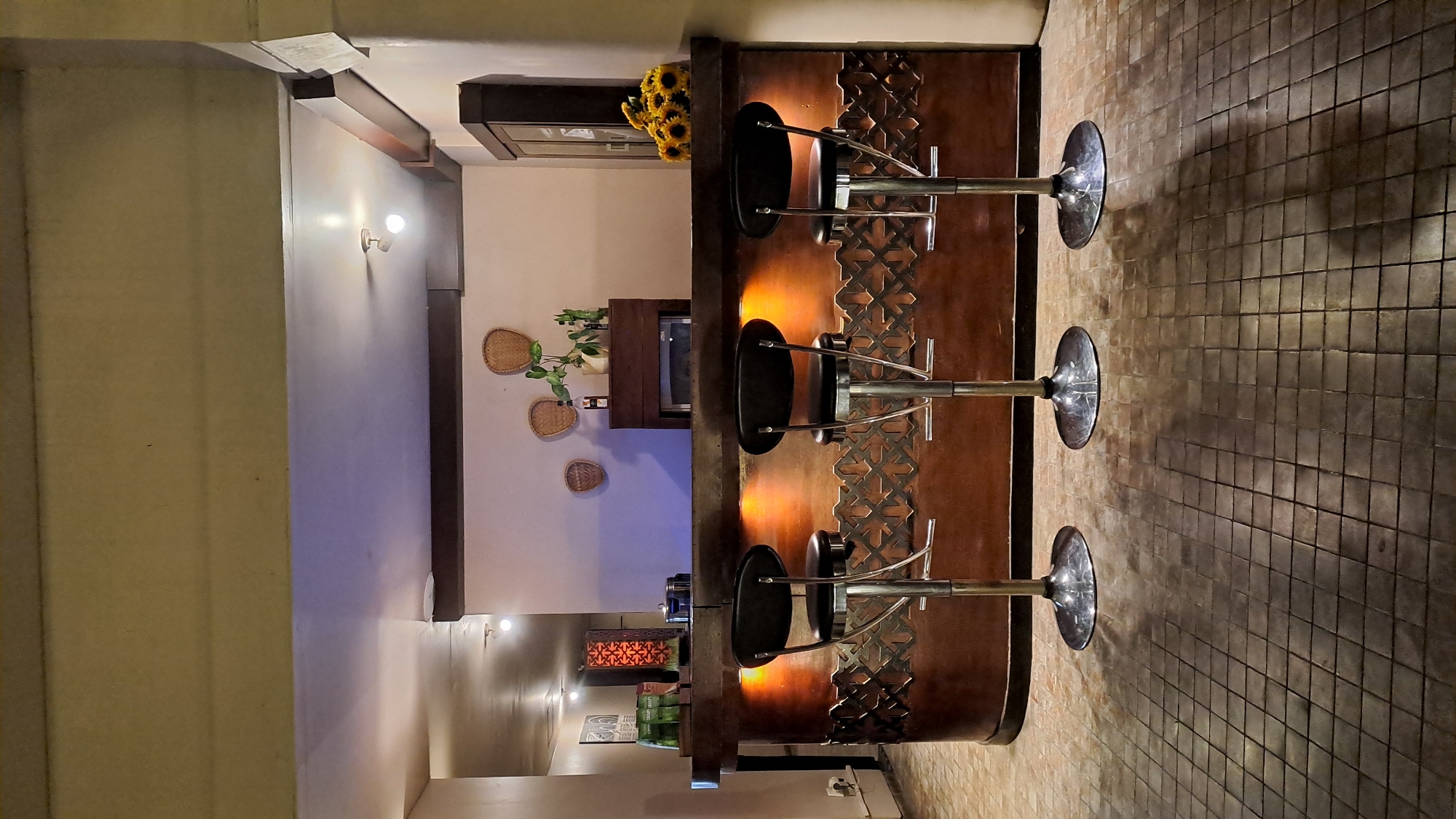 A dimly lit indoor bar scene features a wooden bar with three black stools in the foreground. The bar is illuminated from below, casting a warm orange glow on its decorative front panel. Behind the bar, a dark wooden cabinet stands to the right, adorned with a bouquet of sunflowers. On the wall above the bar, two woven baskets and a small potted plant are visible. The floor is tiled with a mosaic pattern. The overall ambiance is intimate and sophisticated. The location is identified as a bar in Banda Aceh, Indonesia. There are no people present in the image.