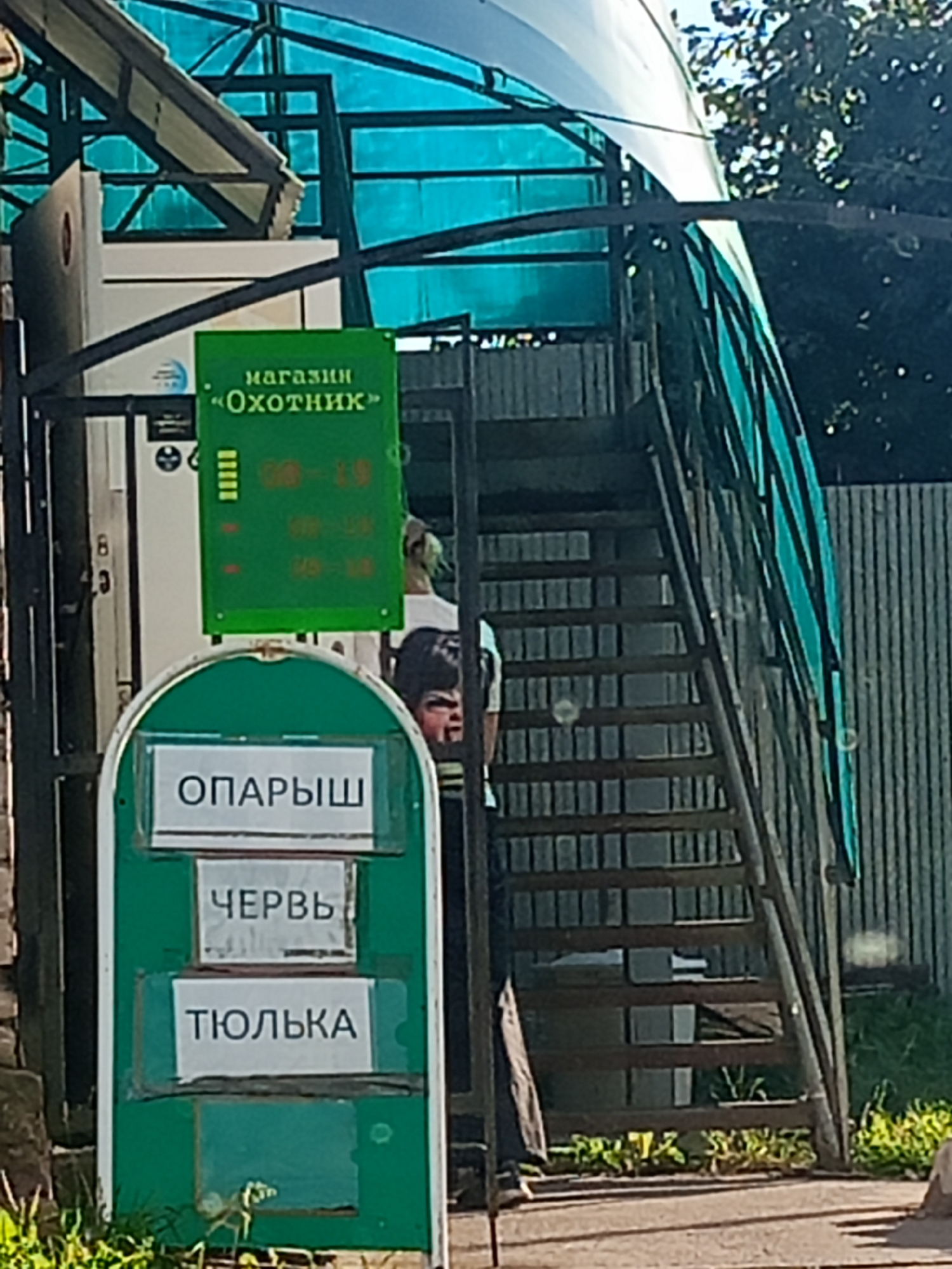 In Chaikovsky, Russia, an outdoor scene features a green metal structure with a transparent blue roof. Attached to the structure is a green sign with "магазин «Охотник»" (Shop "Hunter") and a schedule indicating 9:00-19:00. Below this, another green sign with three white rectangular labels displays "ОПАРЫШ" (Maggots), "ЧЕРВЬ" (Worm), and "ТЮЛЬКА" (Small sprat). To the right, a dark metal staircase ascends, partially obscured by the roof structure. A person with blond hair is visible on the stairs. In the foreground, green foliage grows. A portion of a person's face with dark, styled hair is also visible to the right of the signs.