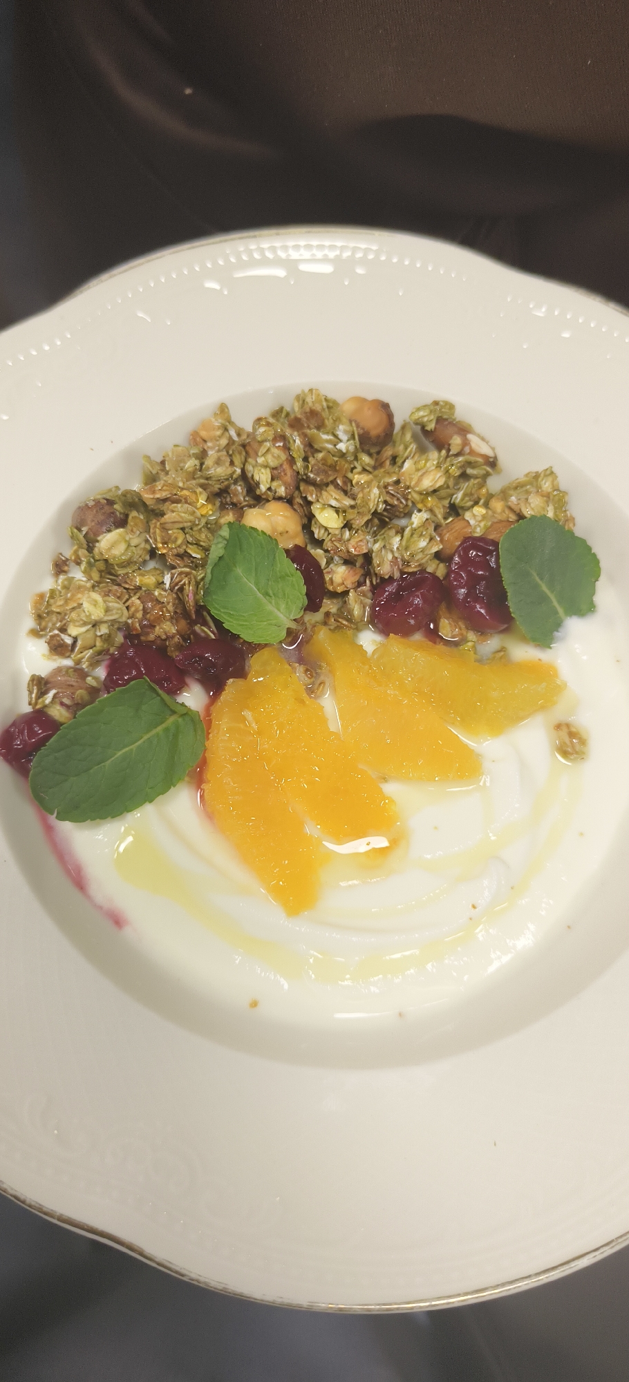 A close-up shot captures a white porcelain plate containing a breakfast or dessert dish. The plate features a decorative edge with a gold trim. Atop a base of creamy yogurt, arranged in a circular swirl, are slices of orange, red berries, and green granola. Three sprigs of mint garnish the dish. A light drizzle of honey or syrup completes the presentation. The background is dark and blurry, suggesting a restaurant or home setting.