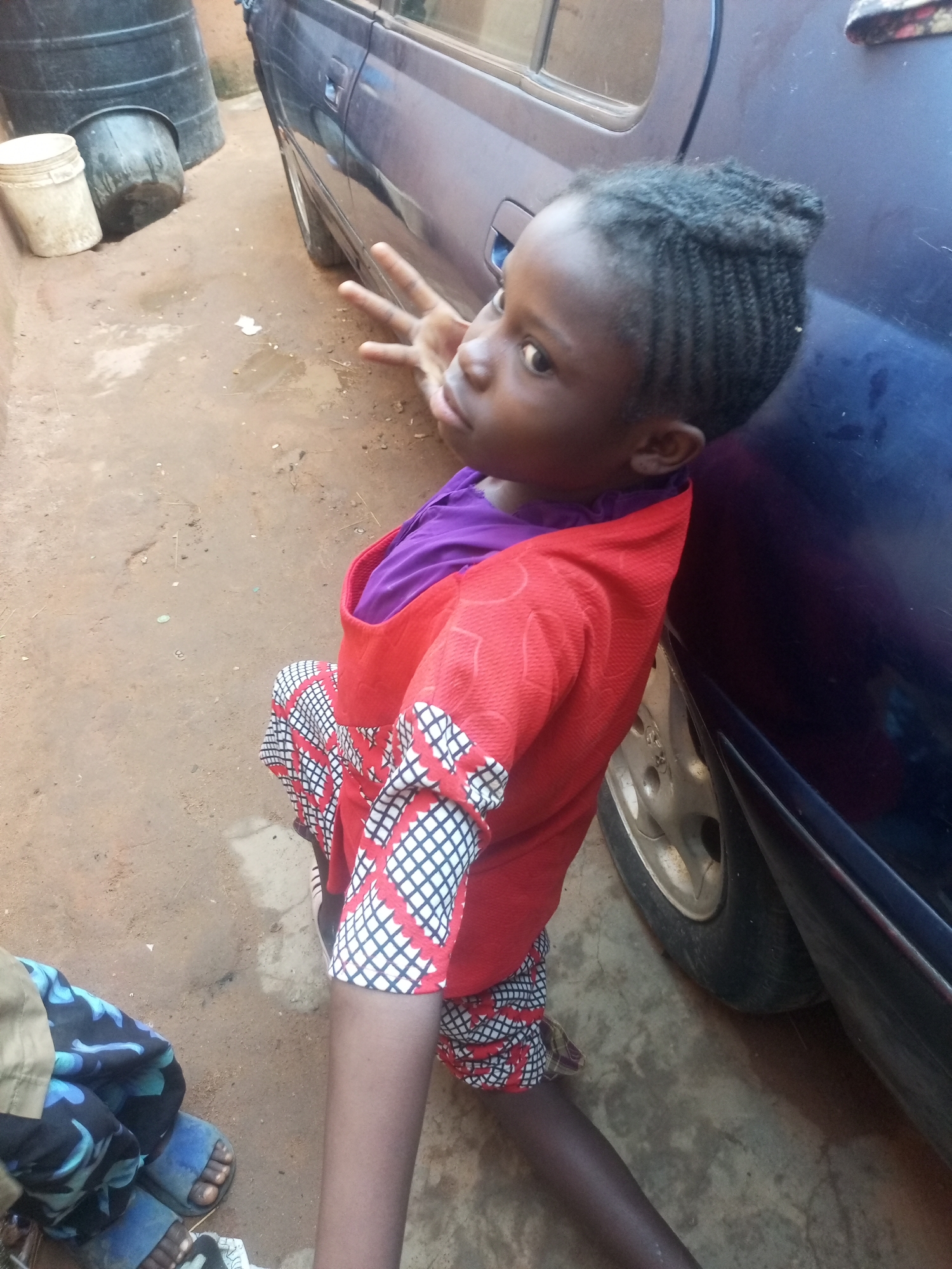 In Tempure, Nigeria, a young Black girl, with braided hair styled in a bun, sits on a dirt ground next to a dark blue car. She is wearing a red garment over a purple shirt, with a skirt or pants featuring a red, white, and black checkered pattern. Her left hand is raised, with fingers extended in a peace sign gesture. The car's wheel is visible, and in the background, large black barrels and a white bucket are situated near a wall. The ground appears to be a mix of dirt and some damp patches, with scattered debris. Another person's legs, wearing blue sandals and patterned pants, are visible in the lower left corner of the image.