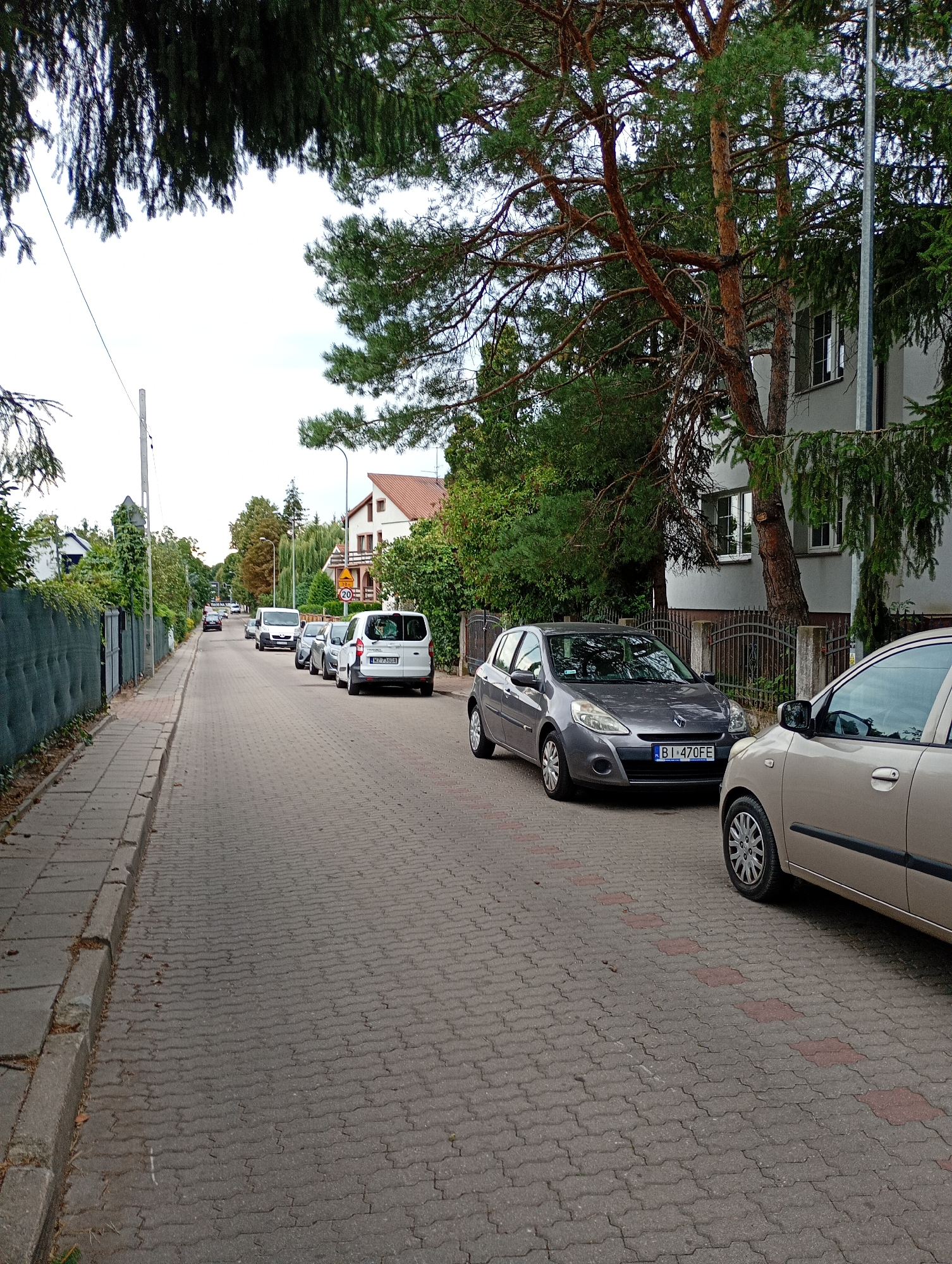 The image shows a street in Białystok, Poland, lined with parked cars and residential buildings. The street is paved with interlocking concrete blocks, and there is a sidewalk on the left side. Trees and greenery are visible along the street, with a tall pine tree prominent in the foreground. Several cars are parked along the right side of the street, including a gray Renault Clio and a white van. A tan car is partially visible in the foreground on the right. A speed limit sign of 30 km/h is visible on the right side of the street. The sky is overcast.