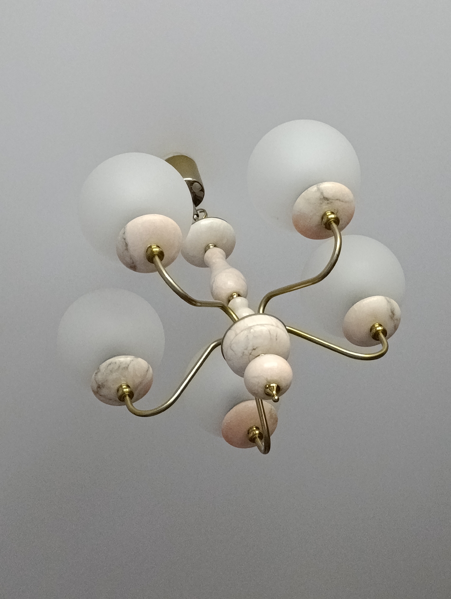 A chandelier hangs from a ceiling. The chandelier has five frosted glass globes, each attached to a brass arm ending in a marble-like disc. The central structure of the chandelier also features marble-like spheres. The ceiling is plain and white. The location is Unknown, Unknown.