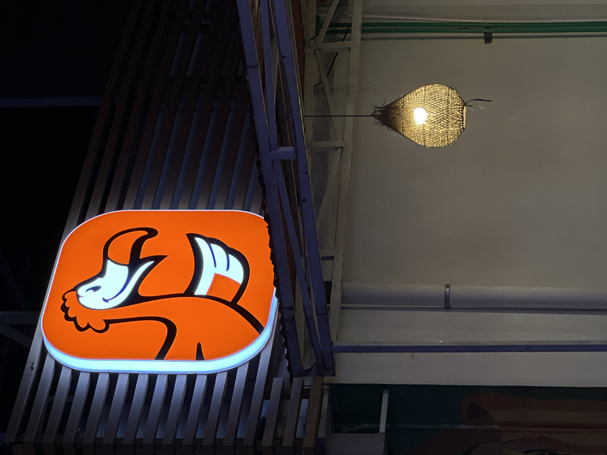 A nighttime shot reveals the orange and white logo of the restaurant "Go! Go! Babi" in North Kuta, Indonesia. The logo, portraying a stylized cartoon pig, is illuminated, and a woven lamp hangs from the ceiling, emitting a warm glow. The building's structure includes a wooden slat design.