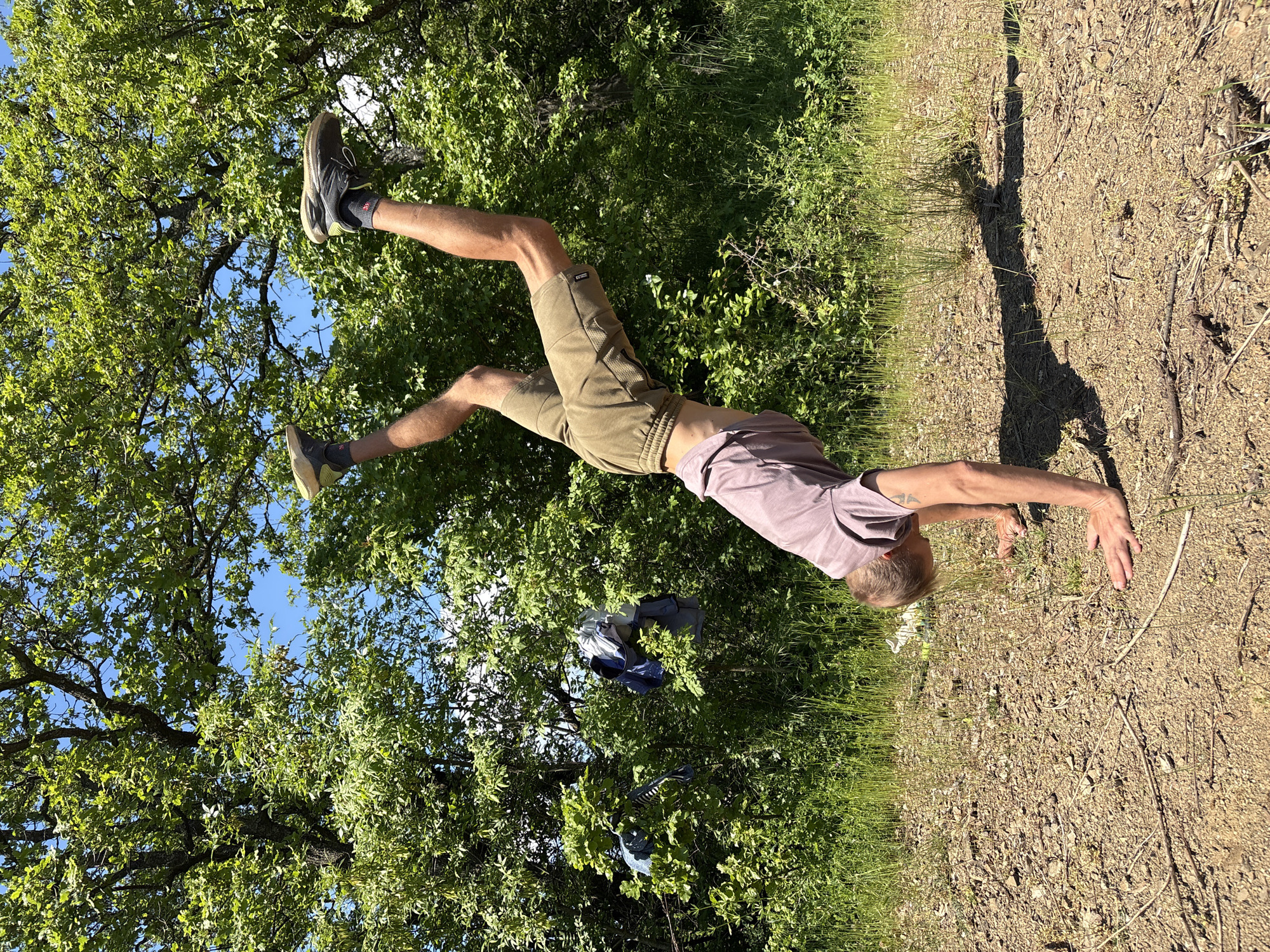 A man is in an inverted position outdoors, with his hands touching the ground, legs elevated towards trees and sky. He wears athletic shoes, shorts, and a t-shirt. The ground is dirt and dry vegetation, transitioning to green grass and bushes. The location is Nizhnee Zaprudnoe, Ukraine. Clothing is caught in the branches of the tree.