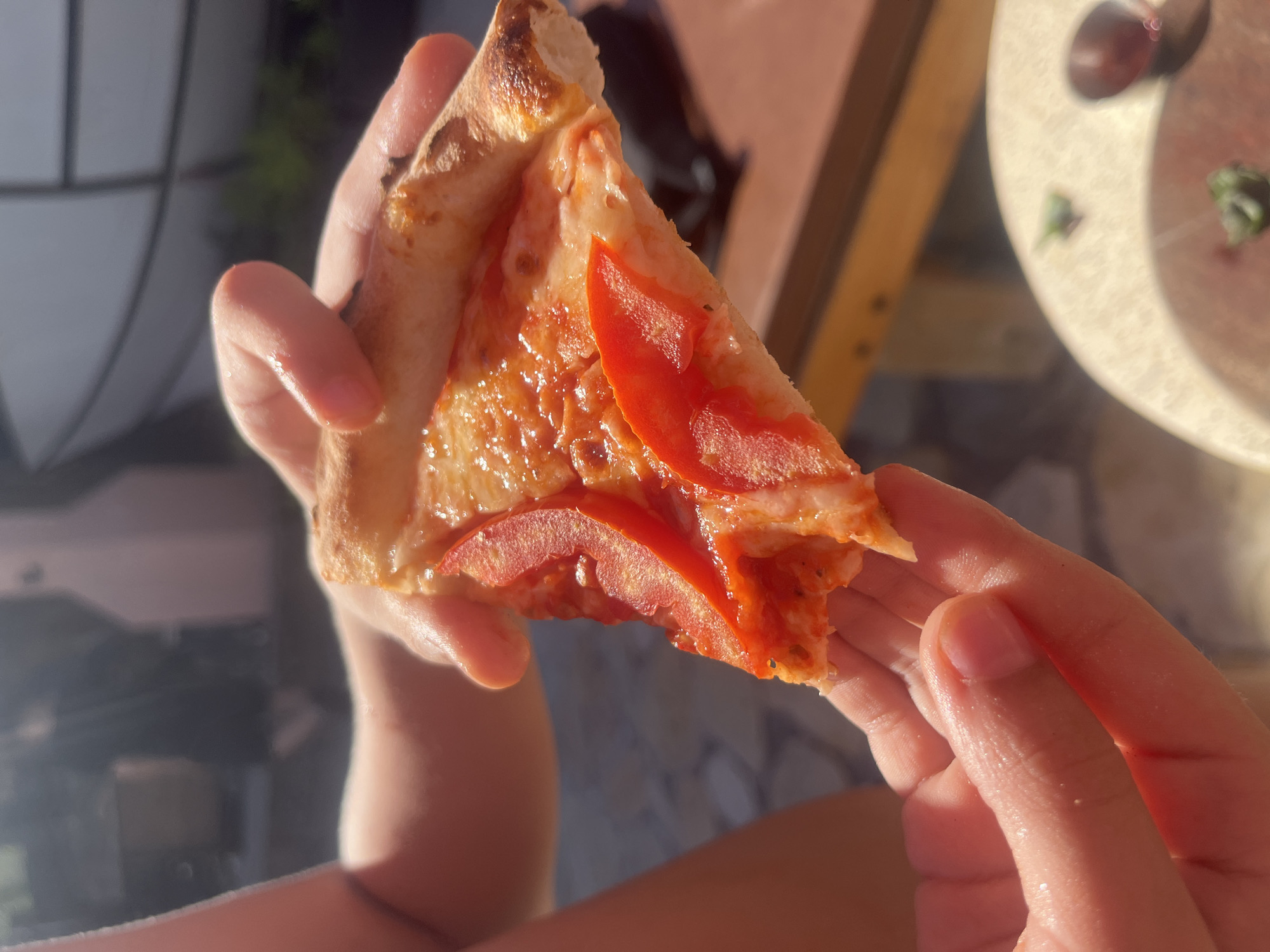 A brightly lit, close-up image captures a person's hands holding a single slice of pizza. The setting appears to be outdoors in Đà Nẵng, Vietnam, bathed in warm, golden sunlight that casts strong highlights on the hands and the pizza's surface.
The pizza slice features a well-baked, slightly charred crust with visible air pockets, characteristic of traditional pizza. It is topped with melted cheese and several vibrant red slices of tomato, which appear juicy and fresh. The surface of the pizza has a glossy sheen, indicating a rich and flavorful preparation.
The person's hands, seen from a high angle, are holding the pizza slice with a gentle grip, suggesting they are about to take a bite or are savoring the moment. Their skin is illuminated by the strong natural light.
The background is softly blurred but hints at an outdoor environment, possibly a patio or balcony. Elements like a light-colored surface (possibly a table) with a faint outline of a glass and some scattered green herbs are discernible, suggesting a casual dining scene. The intense natural light indicates a sunny day, likely during the golden hours of late afternoon or early morning. There is no visible text in the image.