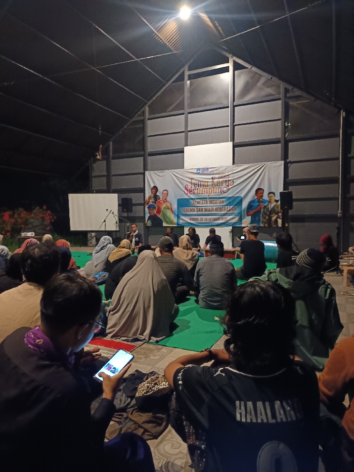The image captures an indoor event at night in Mayang, Indonesia, within a large structure featuring a high, peaked, dark corrugated metal roof and exposed metal support beams. Approximately 20-30 individuals are seated on green mats and a concrete floor, facing a stage area. A single bright light fixture illuminates the overhead space. At the front, a large white banner displays the text "TEMU KARYA SERUMPUNG," "SEMESTA INGATAN TRAUMA DAN IMAJI KEBEBASAN," and "EMBER, 25-28 OKTOBER 2023," accompanied by multiple portrait images. A white projection screen is positioned to the left of the banner. Four to five individuals are present at the stage area, one speaking into a microphone. Two black audio speakers are positioned on either side of the stage. Audience members wear varied attire, some with head coverings. In the mid-foreground, one person is observed using a brightly lit smartphone, while another in the immediate foreground wears a black jersey with "HAALAND" and the number "9" on the back. The scene depicts an organized gathering, likely a seminar or workshop.