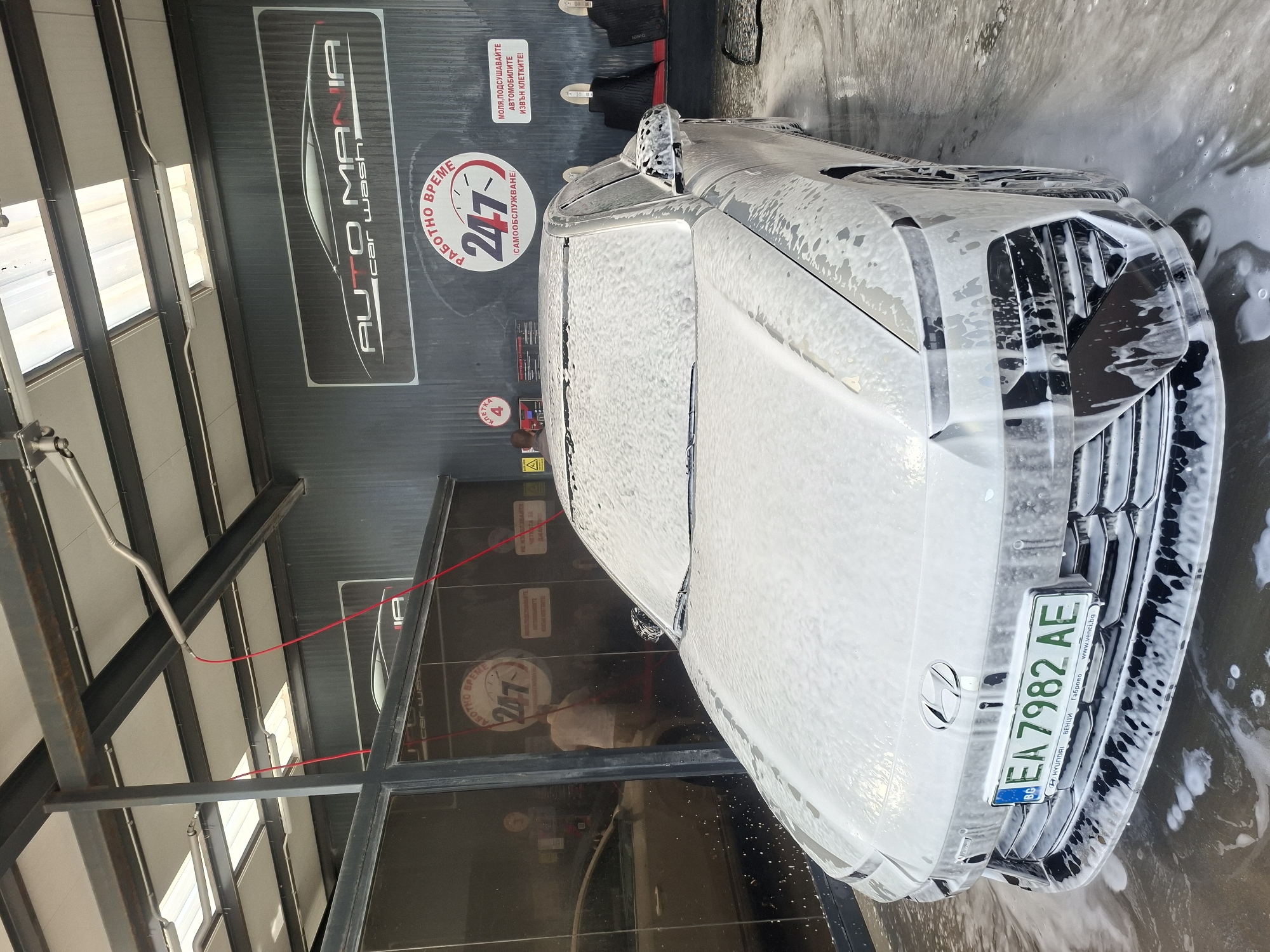 A white Hyundai passenger vehicle is undergoing a car wash at an Auto Mania car wash. The vehicle is covered in soap suds. The license plate number is EA7982 AE. The car wash appears to be located in Габрово (Gabrovo), as evidenced by the “ВЕНЦИ Габрово www.venci.ba” inscription on the license plate frame. The car wash offers self-service, is open 24/7, and customers are instructed to dry their vehicles outside of the stalls.