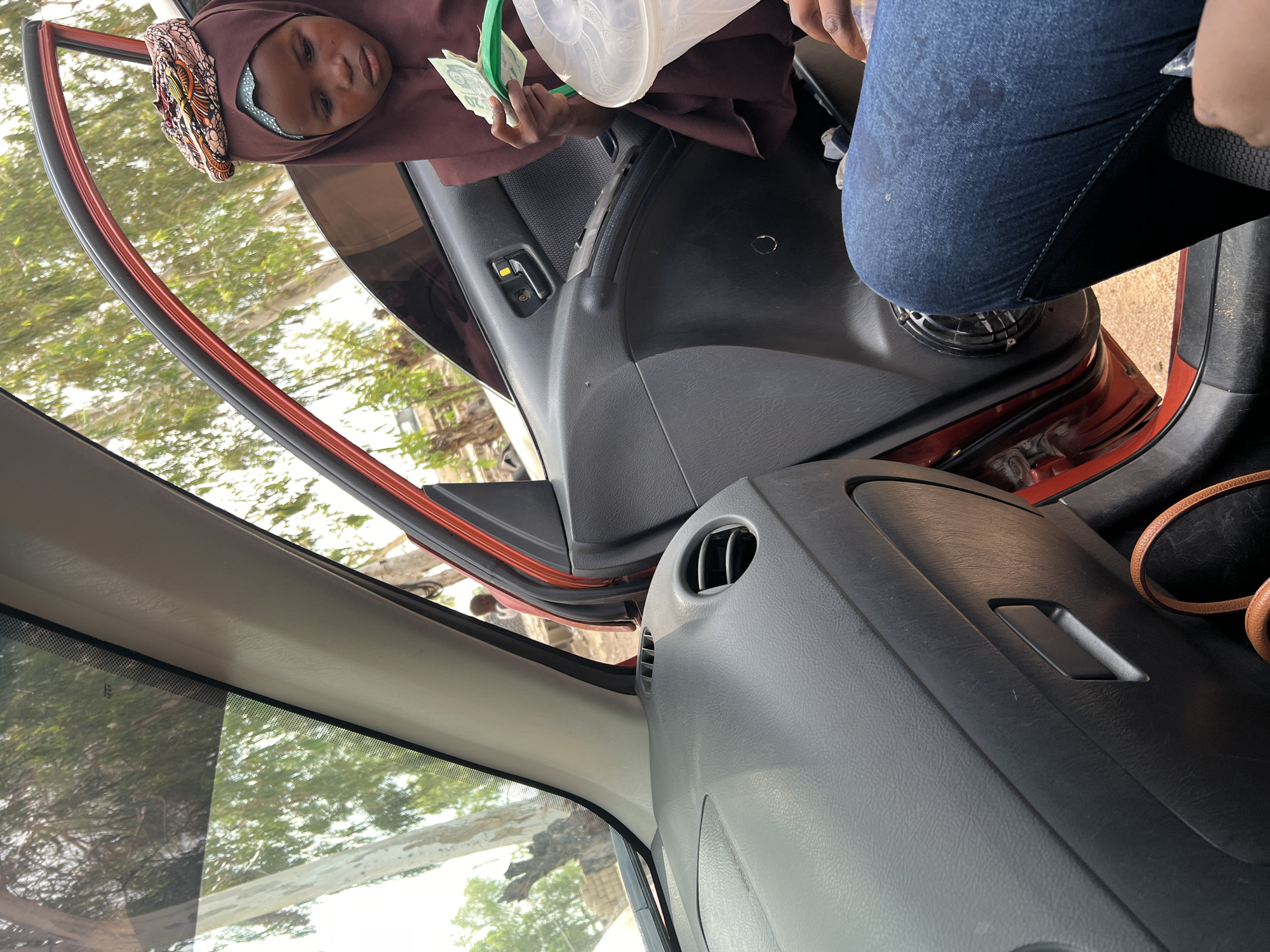 In this image, a young girl wearing a brown hijab with a colorful patterned headband is leaning into the interior of a car. She is holding a clear plastic container and some green banknotes, possibly Nigerian Naira, in her right hand. The car's door is open, revealing the interior, which includes black car door panels, a gray dashboard, and a red door frame. Through the open door and the car's windows, a green, leafy environment is visible, suggesting an outdoor setting. The lighting indicates it is daytime, with sunlight filtering through the trees. The girl's expression is somewhat serious or perhaps contemplative. The scene appears to be in Howayi, Nigeria, as indicated by the provided location context.