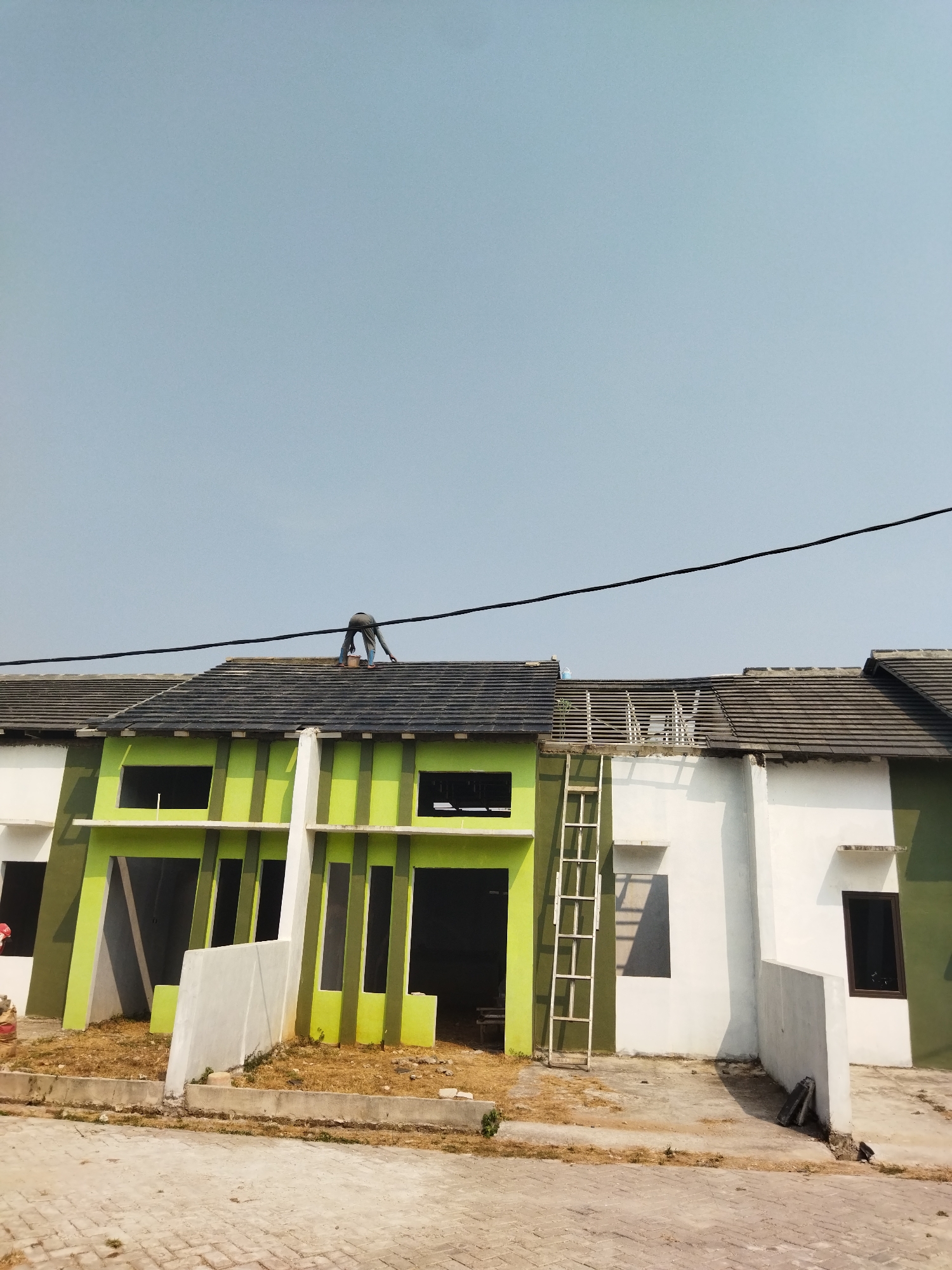 The image shows an exterior view of houses under construction in Suka Sari, Indonesia. A person, dressed in blue pants and a grey shirt, is on the roof of a house, seemingly working. The house has a green and white facade with exposed roof tiles. A ladder leans against the side of one of the houses. The ground in front of the houses is a mix of paved walkways and dry grass. The sky is clear and blue.