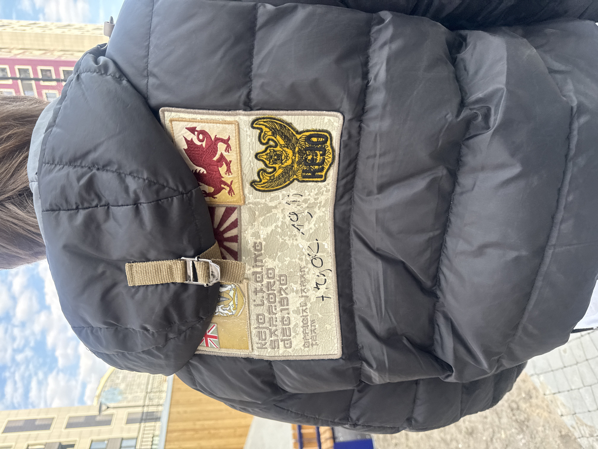 A close-up shot focuses on the back of a black puffer jacket. Affixed to the jacket is a rectangular patch, featuring a grid of various smaller patches and text. The individual patches display a red dragon, a yellow eagle emblem with illegible text, a flag resembling the Rising Sun of Japan, and a British flag. The text on the main patch includes the words "RE-LIKED RED 1970 DEC.1970 OFFICIAL JAPAN". The background is out of focus. In the distance, buildings are visible, which based on architectural styles, suggest a potential location in the city of Saint Petersburg, Russia.