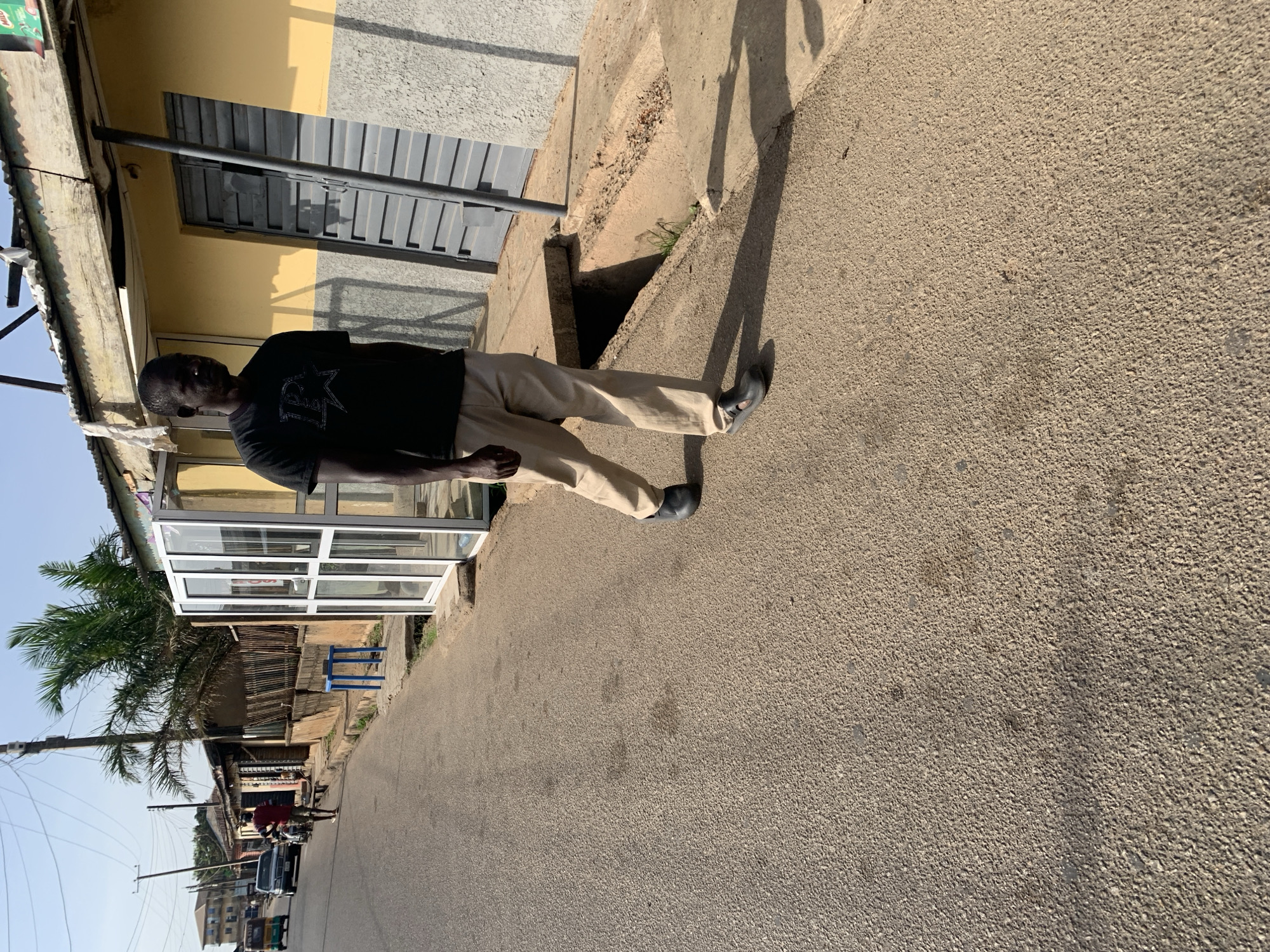The image depicts a street scene in Jos, Nigeria. An adult male stands on the roadside, wearing a black t-shirt and tan pants. He is partially casting a long shadow on the pavement. Adjacent to him is a building with a yellow facade, gray metal shuttered windows, and a white-framed glass storefront. Across the street, a palm tree is visible, along with various buildings and utility poles lining the street. In the distance, cars and a motorcycle are seen traveling down the road. The pavement appears to be rough asphalt. A drainage ditch is present at the edge of the pavement near the building.