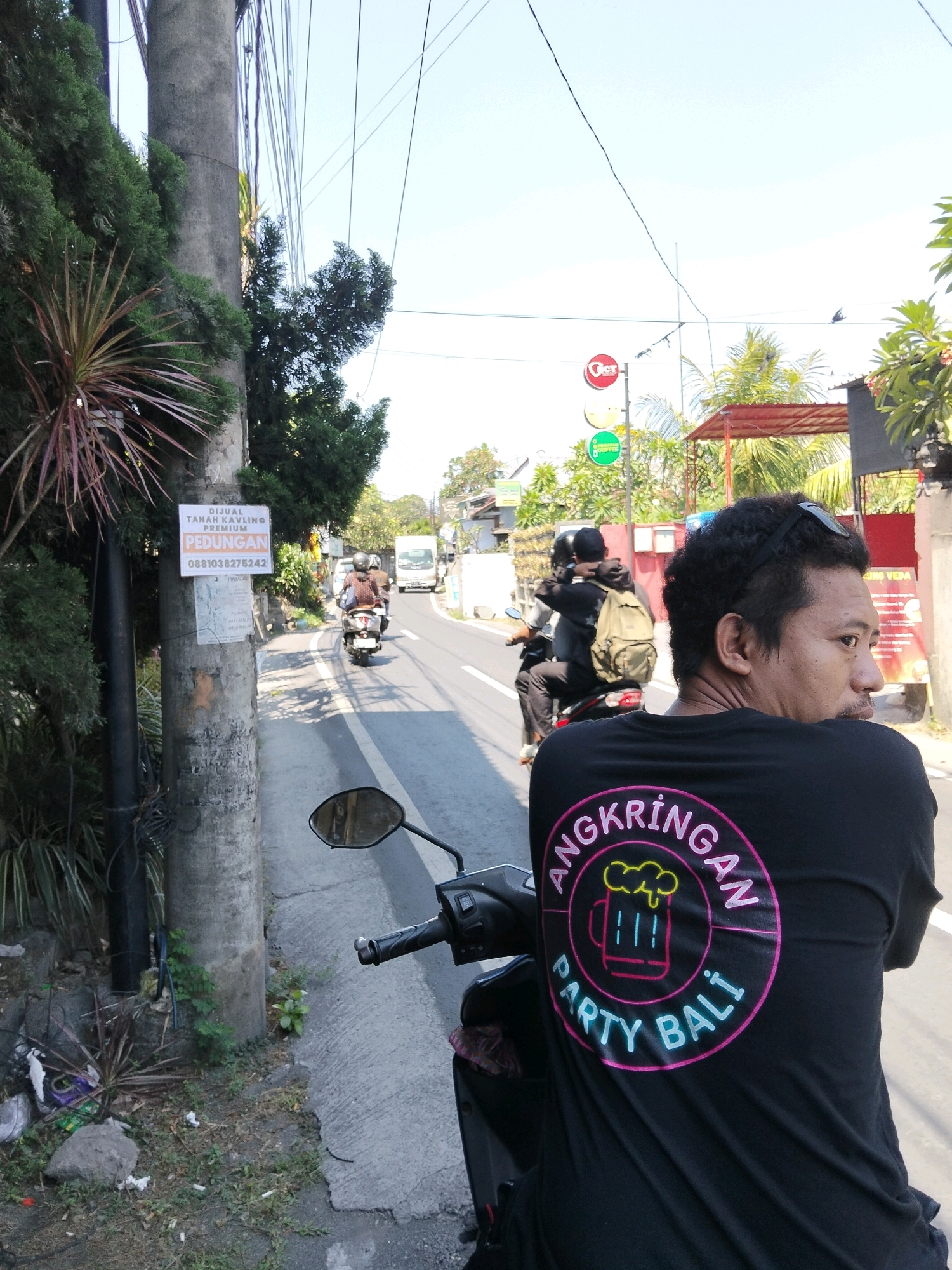 In Denpasar, Indonesia, on a paved road, a man wearing a black t-shirt with a circular graphic featuring a beer mug and the text "ANGKRINGAN PARTY BALI" is riding a motorcycle. His head is turned to the right, looking away from the camera. Behind him, another motorcycle carries two people, one with a backpack. In the background, a white van is visible, along with buildings, palm trees, and electrical wires overhead. A utility pole on the left side of the road has posters attached to it, including one that appears to be an advertisement for land sales. The scene is illuminated by daylight, suggesting it is daytime.