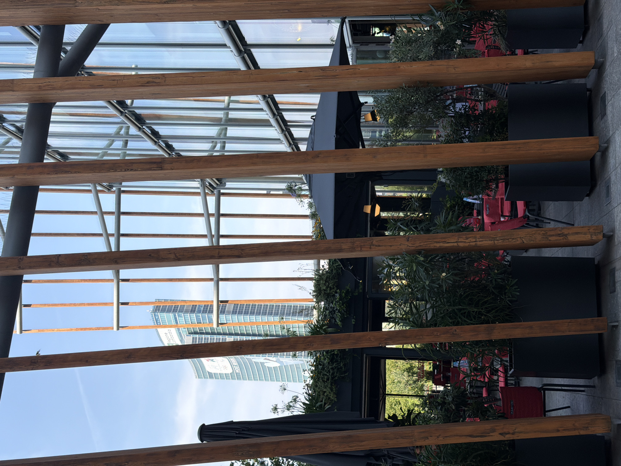 The image depicts an outdoor seating area of a cafe or restaurant in Milan, Italy, with large wooden beams in the foreground obscuring the view. Behind the beams, a black umbrella is visible, along with several red chairs and tables set up for dining. Planters with lush greenery are placed around the seating area. In the background, two modern glass skyscrapers rise into a clear blue sky, one featuring a prominent advertisement for "Regione Lombardia". The architecture suggests a contemporary urban setting within Milan. There are no people visible in the image.