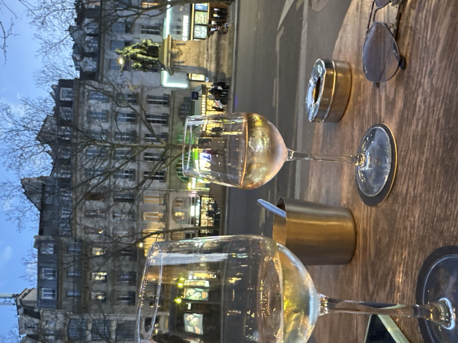 The scene depicts an outdoor café table at dusk in Paris, France, specifically on the Quai de l'Hôtel de Ville, facing the equestrian statue of Étienne Marcel. In the immediate foreground, a wooden table holds two stemmed glasses, each containing a pale yellow liquid. The right glass is fully visible, resting on a dark coaster, while the left glass is partially cropped. Adjacent to the glasses are a small, brushed metal cylindrical holder with a spoon inserted, a silver-toned oval ashtray with an indented lid, and a pair of dark aviator sunglasses. A portion of a mobile phone is visible at the bottom left.
Beyond the table, an asphalt road runs left-to-right, separating the viewer from the opposite sidewalk. Pedestrians are visible walking on this sidewalk. Commercial establishments with illuminated signs, including one partially displaying "BRASSERIE," line the street. Several individuals are seated at outdoor tables on the opposing side. To the right, a large bronze equestrian statue on a stone pedestal is prominent, depicting a rider and a standing figure. This monument is the Statue of Étienne Marcel. The background consists of classic Parisian Haussmannian buildings, characterized by multiple stories, stone facades, and mansard roofs, with numerous windows illuminated. Bare deciduous trees are interspersed along the street and in front of the buildings. The sky above is a deep twilight blue, indicating the transition from day to night.
