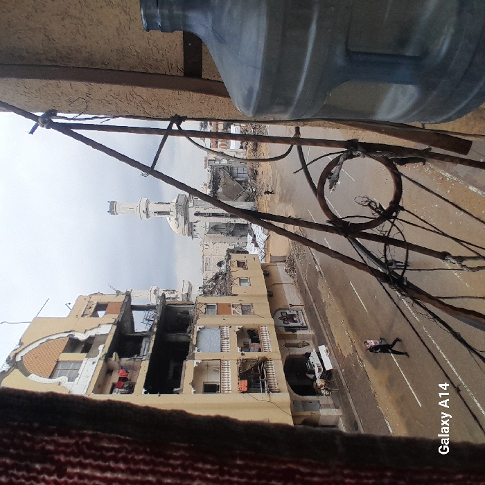 An aerial perspective of a street in Khan Yunis, Palestinian Territory, reveals a scene of significant urban destruction under an overcast sky. In the immediate foreground, a reddish-brown textured fabric partially frames the left edge, while a large, translucent blue plastic water carboy and a tan building wall frame the right. A rusted metal railing with circular decorative elements and attached dark wires extends diagonally across the right foreground, partially obscuring the view below.
To the left, a multi-story, light-colored building displays severe structural damage, with multiple floors exposed and internal elements visible. Clothes are seen hanging on a line from a lower-level balcony of this damaged structure. The ground level includes what appear to be partially destroyed commercial facades.
A wet, asphalt road with white lane markings spans the midground. One individual, dressed in dark clothing, walks rightward on this street, appearing to carry a light-colored object or small person on their back. A smaller, darker figure is discernible further down the street.
The background is dominated by a large, light-colored mosque featuring a central dome and two minarets. The mosque exhibits considerable damage, with visible collapse of sections of its dome and surrounding structures. Extensive piles of rubble and debris are scattered across the areas surrounding the mosque and lining the street. A solitary dark figure is observed standing amidst the debris in the distance near the mosque.