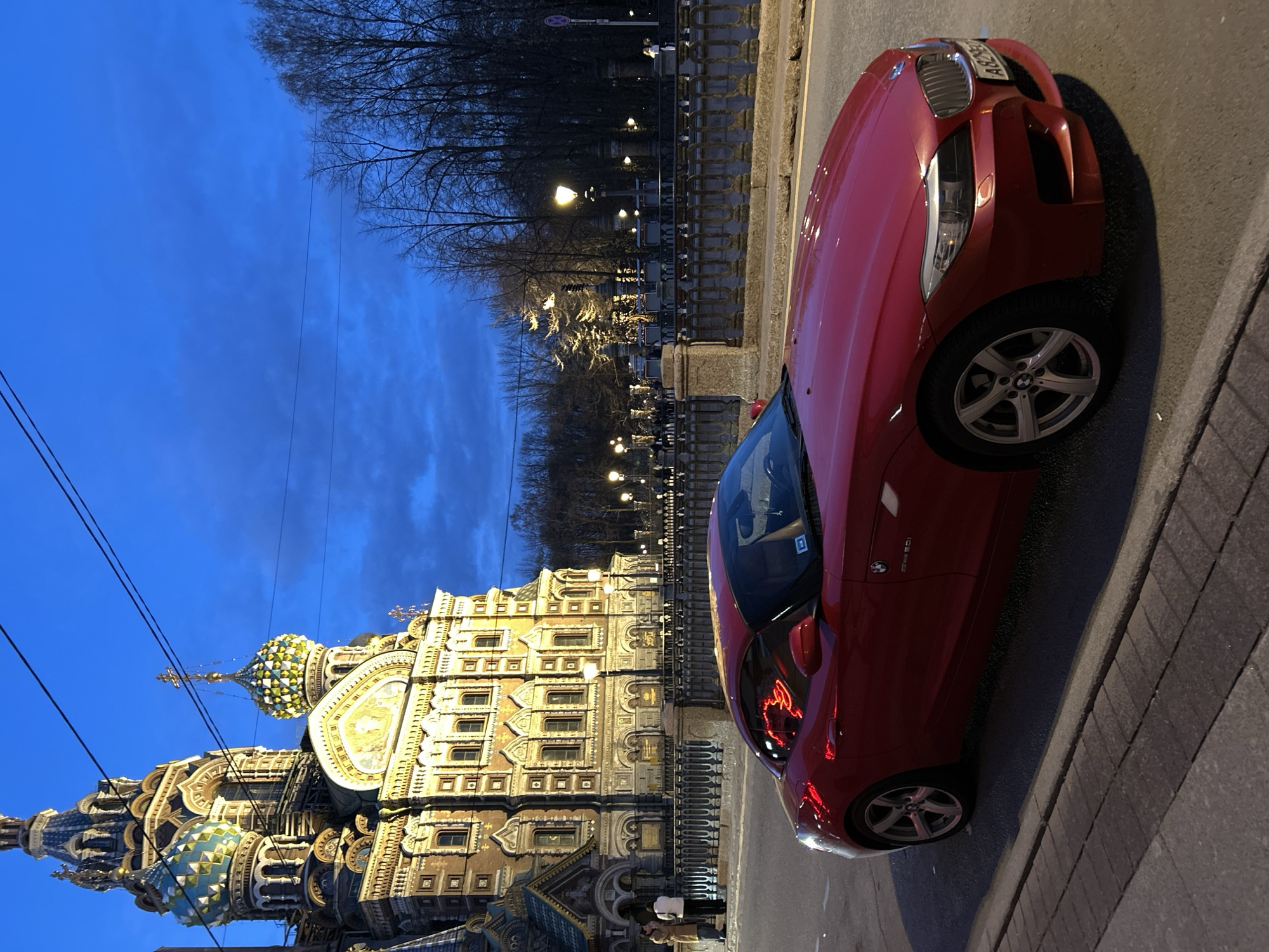 The image captures a vibrant twilight or early evening scene in Saint Petersburg, Russia, with the iconic Church of the Savior on Spilled Blood dominating the background. The sky is a deep, rich blue, indicating the transition from dusk to night, with streetlights and architectural lighting illuminating the surroundings.
In the foreground, a glossy red BMW Z4 sports car is parked on the right side of the street. Details like the "Z4" badge and BMW emblem on the wheels are visible, along with a partial Russian license plate that begins with "A82 BC". The car casts a soft shadow on the pavement, and reflections of the city lights, including what appears to be neon, are visible on its body and windows.
On the left, the magnificent Church of the Savior on Spilled Blood stands majestically. Its elaborate facade, adorned with intricate mosaics and carvings, glows warmly under artificial illumination. Several colorful, onion-shaped domes, characteristic of Russian Orthodox architecture, are prominent against the darkening sky.
Between the car and the church, a sidewalk runs parallel to a decorative iron fence. Beyond the fence, a line of bare trees suggests either late autumn, winter, or early spring. Scattered streetlights cast pools of light along the street and sidewalk. Two small figures, appearing to be people, are visible near the base of the church, adding a sense of scale to the grand structure. Overhead, thin power lines stretch across the frame. The overall scene is calm and atmospheric, highlighting the beauty of the historic landmark juxtaposed with modern urban elements.