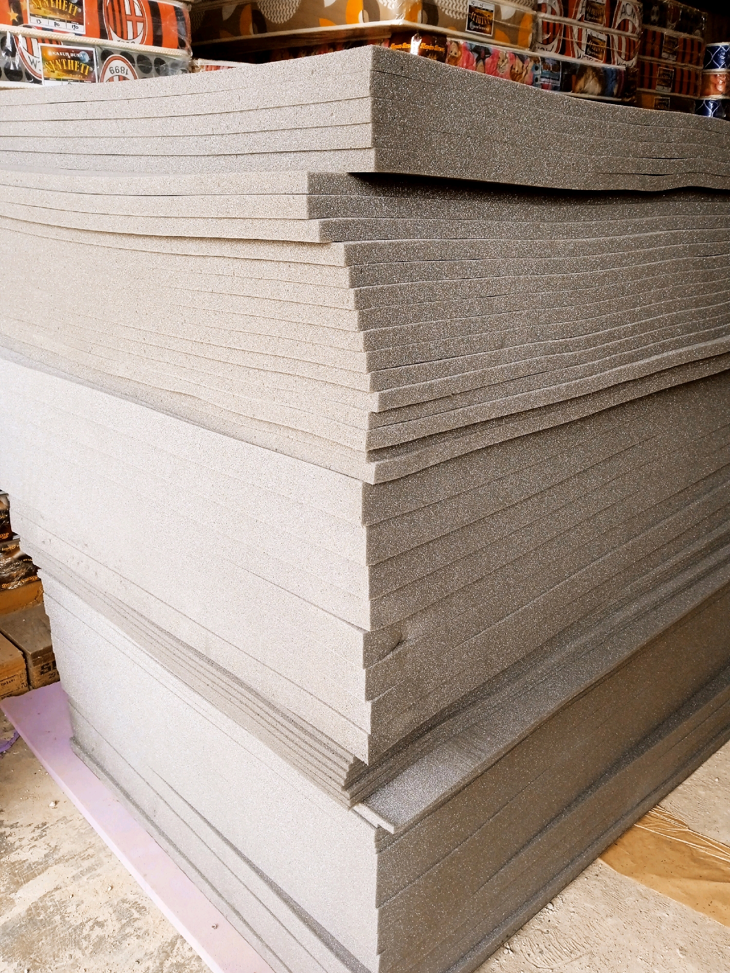 A stack of foam sheets is pictured in a warehouse or storage area in Banjarbaru, Indonesia. The sheets are of varying colors, with some appearing light gray and others a darker gray or brown. They are stacked neatly on top of each other, creating distinct layers that are visible due to the lighting and the texture of the foam. In the background, there are some boxed items, possibly mattresses or other packaged goods, also stacked. The floor appears to be concrete and dusty, with some other materials visible, including a pink sheet of foam underneath the main stack. The overall impression is one of a commercial or industrial storage space.