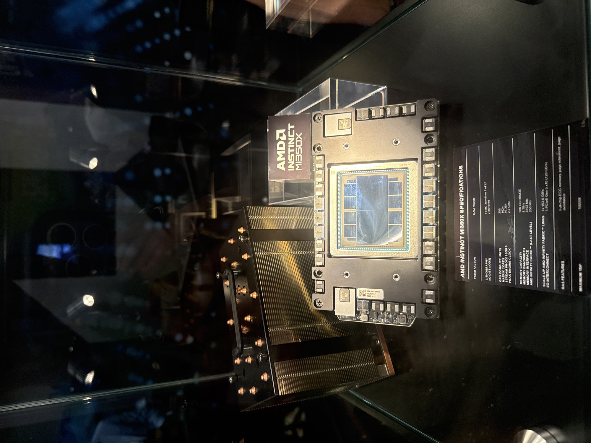 This image depicts an AMD Instinct MI350X accelerator, displayed in a glass case. A metal heatsink is attached to the left. A card with specifications is visible, listing details like "288 GB HBM3E" memory and "256 GPU Compute Units." The reflection suggests the photo was taken in a public space, possibly a tech conference or exhibit. There is no indication of the image being taken in Vancouver, Canada.