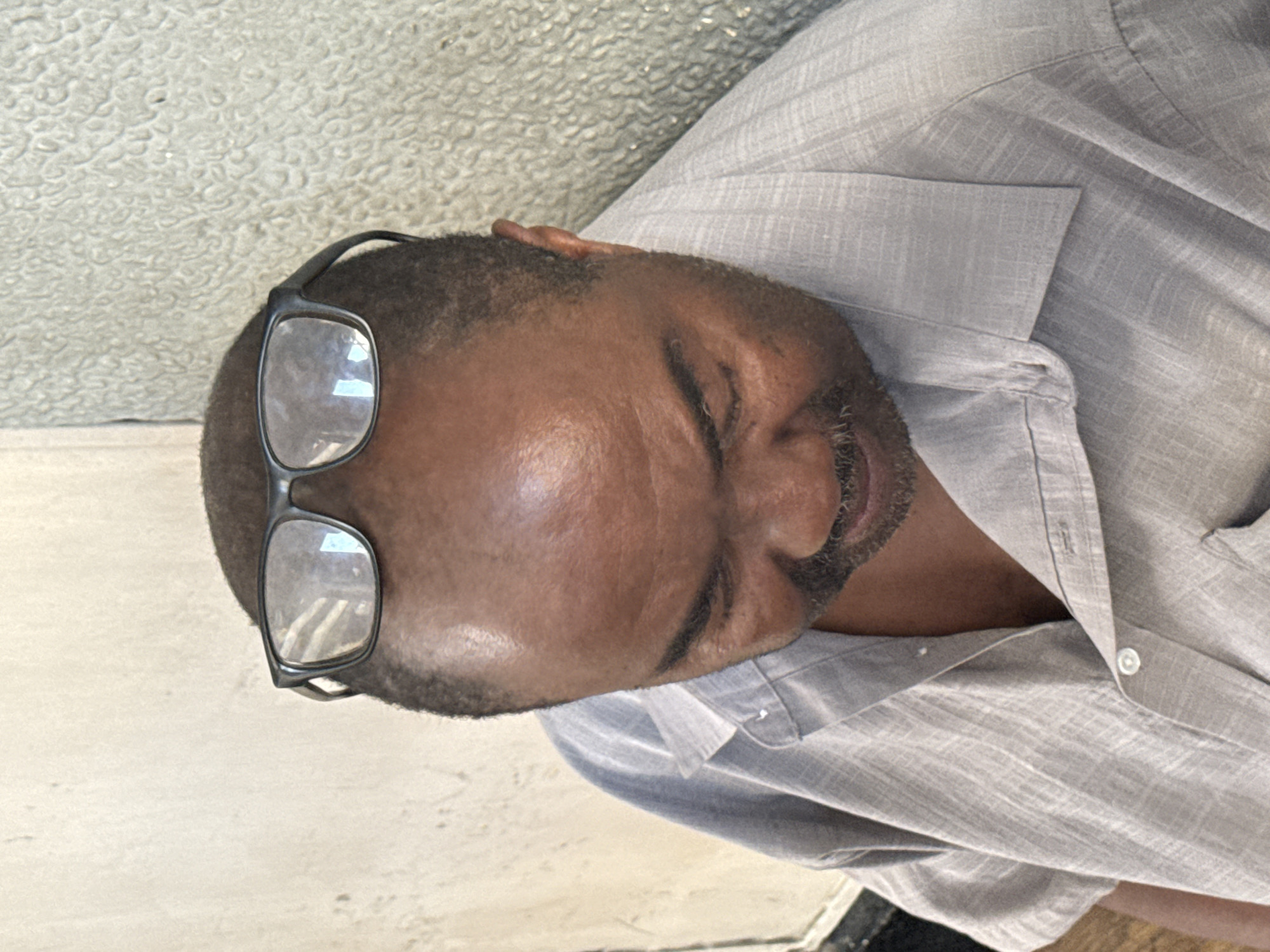 A medium shot captures a Black man wearing glasses perched on his head, facing downwards and to the right. He has a receding hairline, dark skin, and a mustache and beard. He is wearing a light gray collared shirt with short sleeves and buttons. The background consists of a textured, light-colored wall to the left and a rough, mottled gray surface to the right. The scene appears to be an indoor setting, possibly a waiting area or a simple room. There is no specific indication of the location within Dar es Salaam, Tanzania, beyond the general description of the environment.