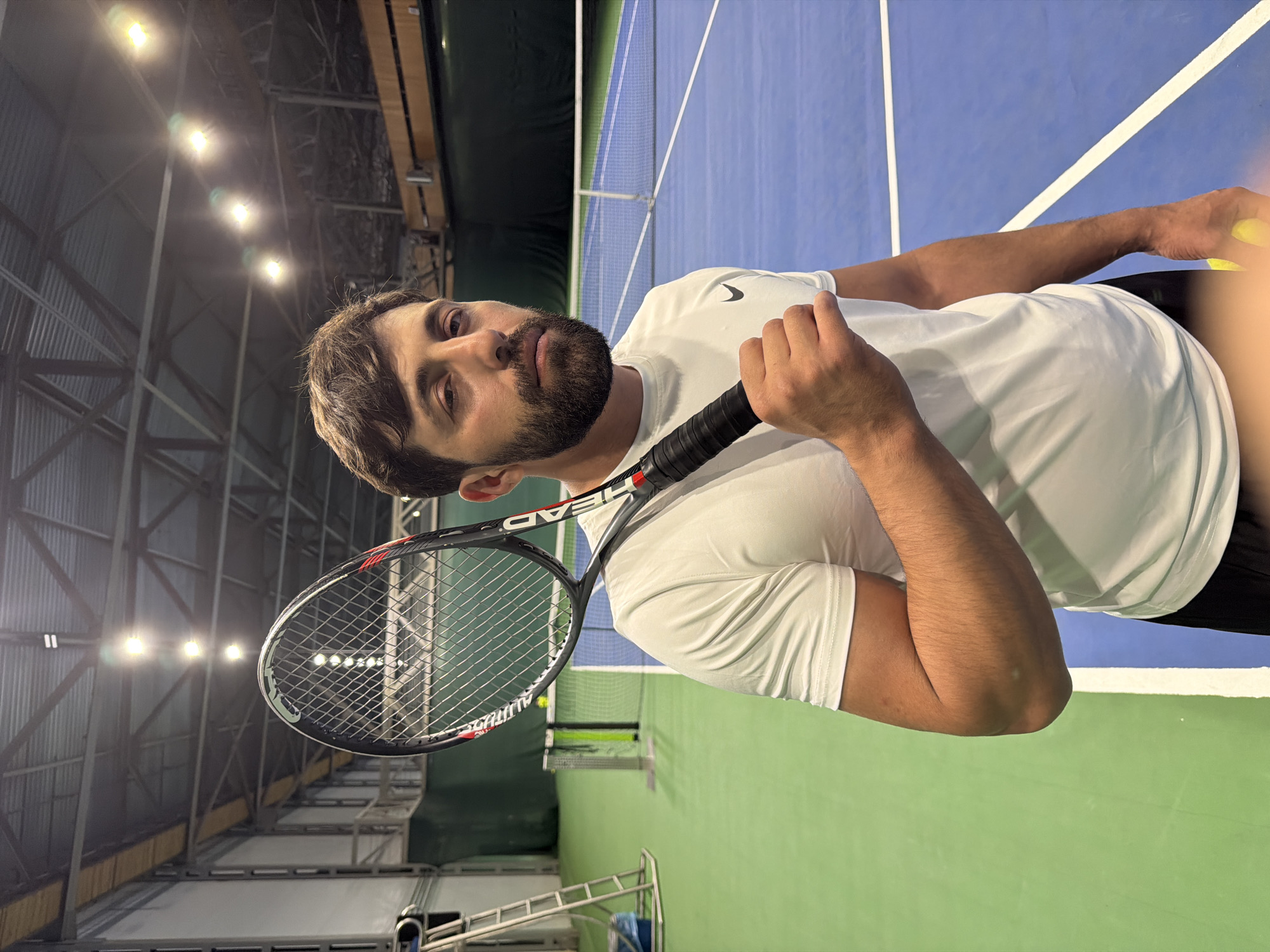 A medium shot captures a man on a tennis court, possibly indoors, with the camera rotated approximately 90 degrees. He is holding a tennis racket in his left hand and a tennis ball in his right. He is wearing a white t-shirt and black shorts. The tennis court is divided into green and blue sections, separated by a net. The location is likely Krasnoyarsk, Russia, due to its geographical location, the architecture of the indoor tennis facilities visible in the background, and the likelihood of this activity occurring in a Russian city. Overhead are industrial-style lights and structural elements.
