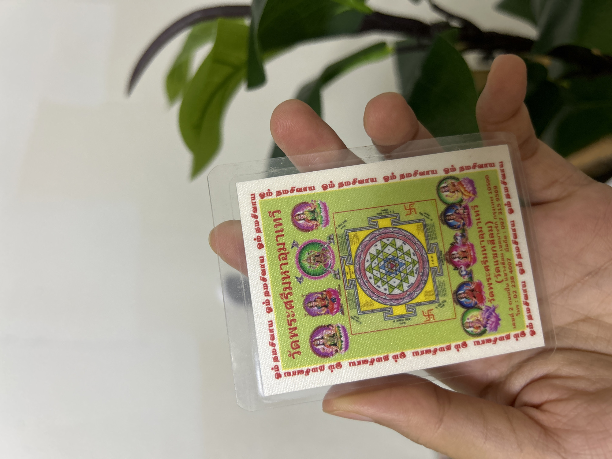 A person is holding a laminated card with religious imagery. The card features a central mandala-like design surrounded by depictions of deities and decorative borders. The text on the card is in Thai. Below the main design, there is an address: "เลขที่ 2 ถนนปิ่นเกล้า แขวงบรมราชชนนี เขตตลิ่งชัน กรุงเทพฯ 10500" and a phone number "โทร : 02 238 4007, 097 315 9569". These details indicate that the card's origin or the depicted temple is located in Bangkok, Thailand, not Khlong Luang Town Municipality. The background is blurred and shows green leaves of a plant, suggesting an indoor setting. The lighting is bright, illuminating the card.