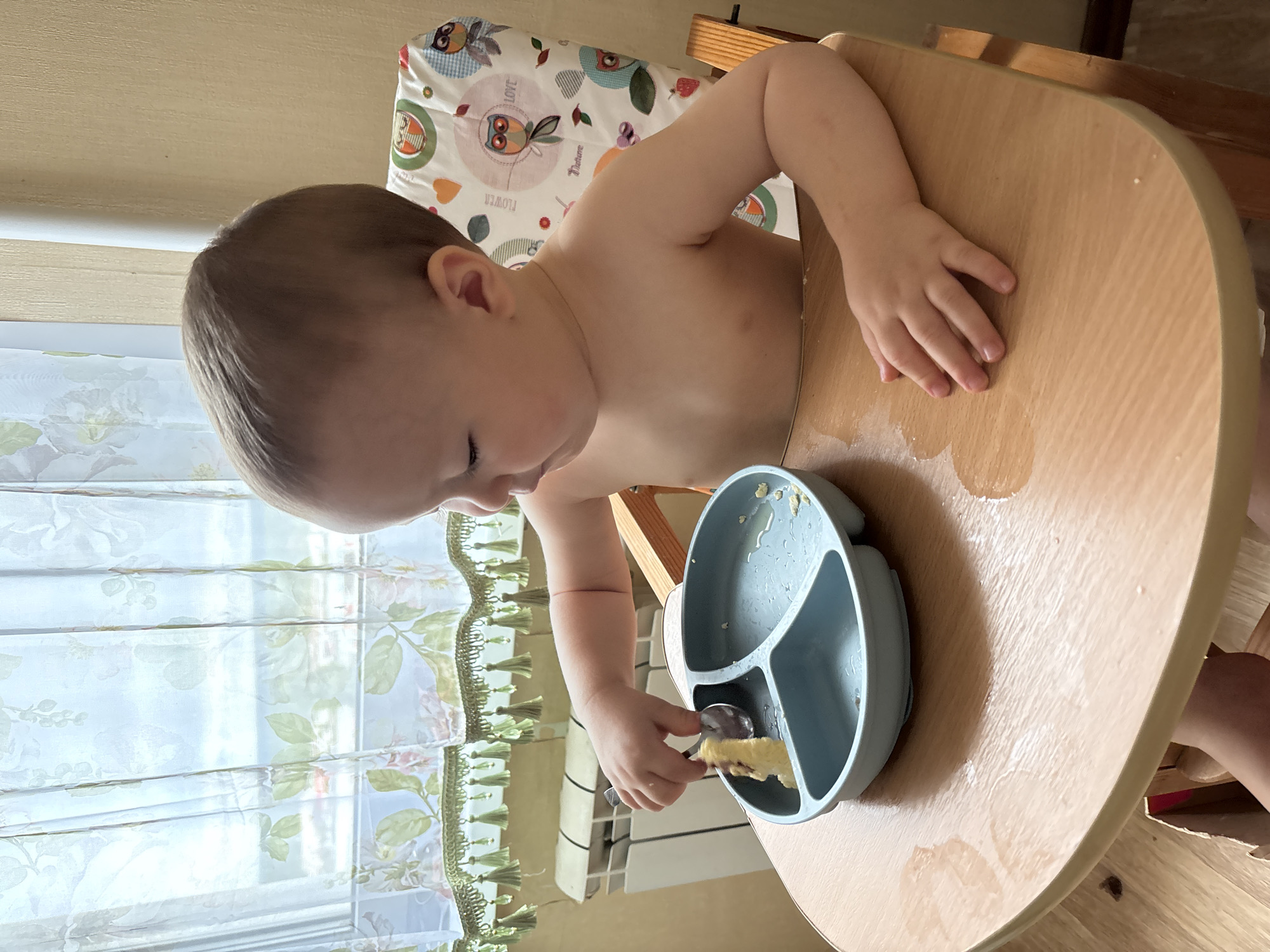 An overhead, medium shot captures a toddler sitting in a wooden high chair, facing right, with their upper body exposed. They are holding a spoon in their right hand, attempting to pick up a piece of food from a partitioned blue plate placed on the wooden tray of the high chair. The tray is wet with spilled liquid, indicating a meal in progress. Behind the child, sheer curtains with floral patterns and green fringe cover a window, through which daylight is visible. A white radiator is situated beneath the window. The high chair has a colorful fabric backing with illustrations of owls and other motifs. The scene appears to be indoors, possibly a dining area in a residential setting in Naberezhnye Chelny, Russia, given the context provided.