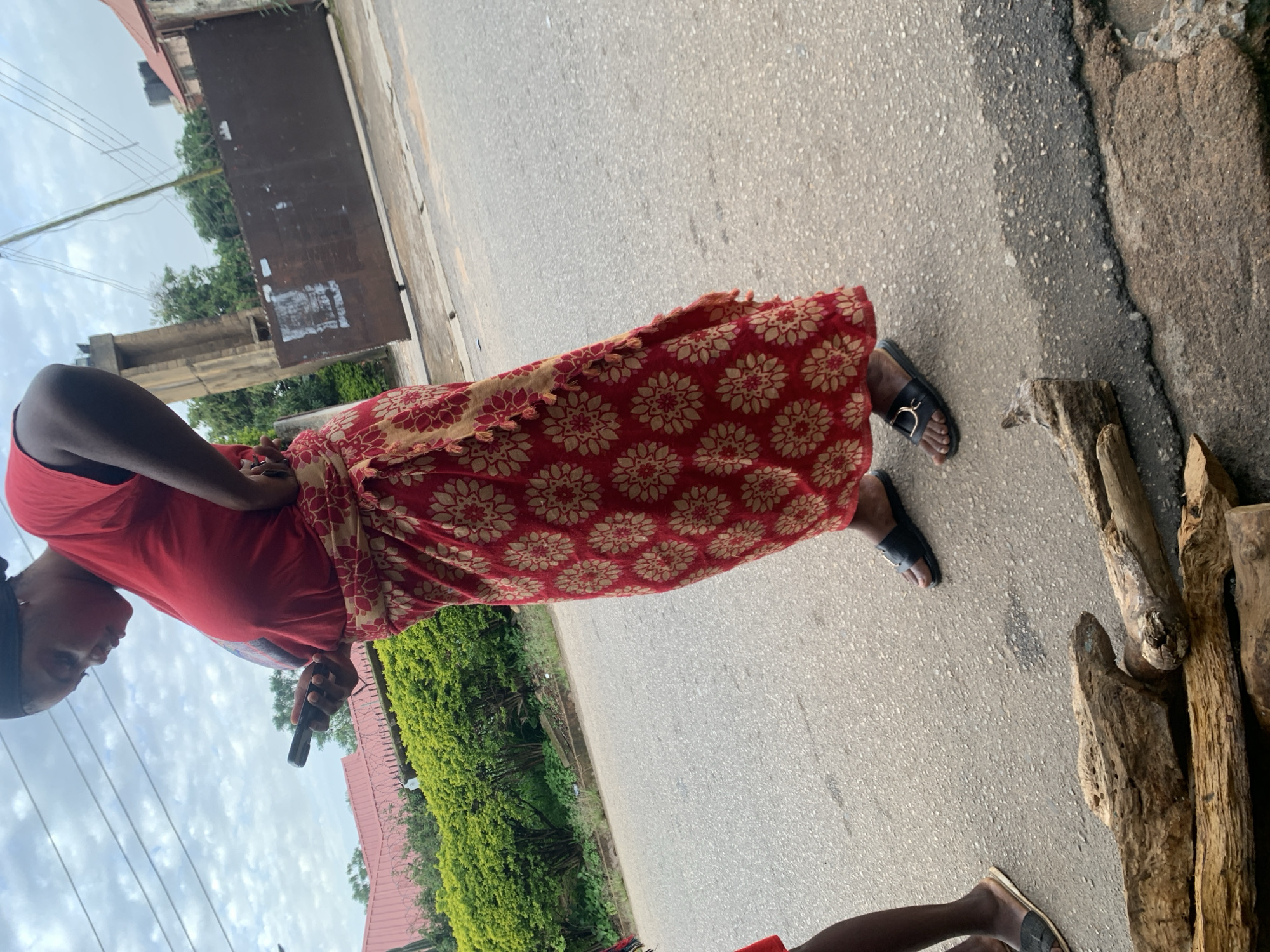 An overhead shot of a woman standing on a street in Jos, Nigeria. She is wearing a red t-shirt, a red and gold patterned skirt, and black sandals. She is holding a black smartphone in her right hand and looking down at it. In the foreground, several pieces of wood are scattered on the asphalt. Behind the woman, a hedge and a rusted metal gate are visible. The sky is overcast.