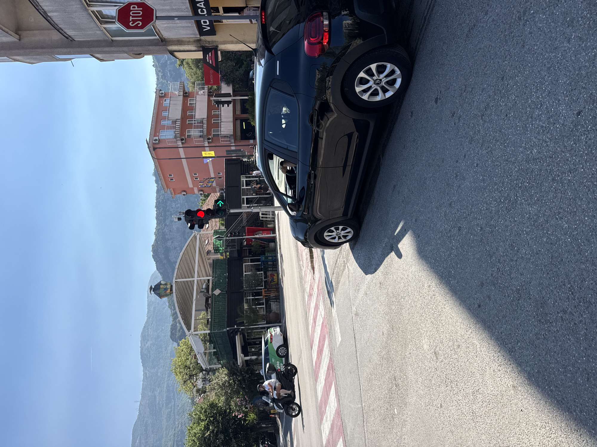In Budva, Montenegro, this daytime street scene captures a busy intersection under a clear blue sky. A black four-door hatchback, identified as a Citroën C3, is stopped at a pedestrian crossing, its rear brake lights illuminated. The car's driver is visible through the window. Adjacent to the car, a traffic light displays a red signal for one direction and a green signal for another.
Across the street, a man on a three-wheeled scooter is navigating the intersection. In the background, a two-story building with a pink facade stands tall, featuring multiple balconies with white railings and air conditioning units. To its right, a taller, gray building displays a red octagonal "STOP" sign. Further in the distance, lush green mountains form a picturesque backdrop.
The area features businesses with awnings, including one with a "VO.CAME" sign. A pedestrian crossing with red and white stripes is clearly marked on the asphalt. The ground is paved with dark asphalt, and shadows are cast by the sunlight. Trees with green foliage are scattered throughout the scene, adding to the urban landscape.