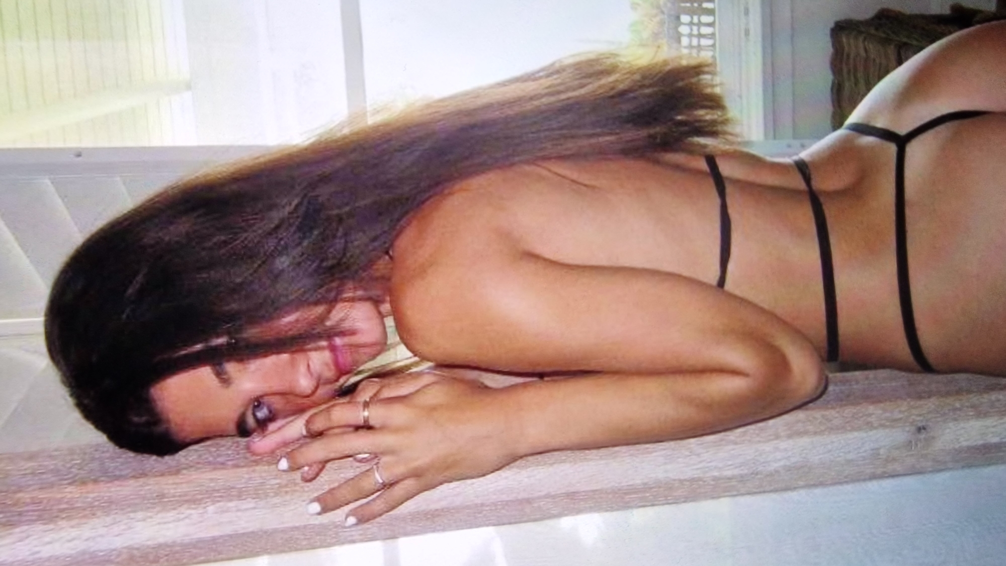 The image shows a woman wearing a black bikini. She has long, dark brown hair and is looking over her shoulder at the camera. She is standing next to a beige curtain, and in the background, there is a window with white blinds. There are some plants visible outside the window, and a woven chair can be seen to the right. The scene appears to be indoors, possibly in a home or hotel room. The woman has her right arm bent and her hand near her face. She is wearing rings on her fingers.