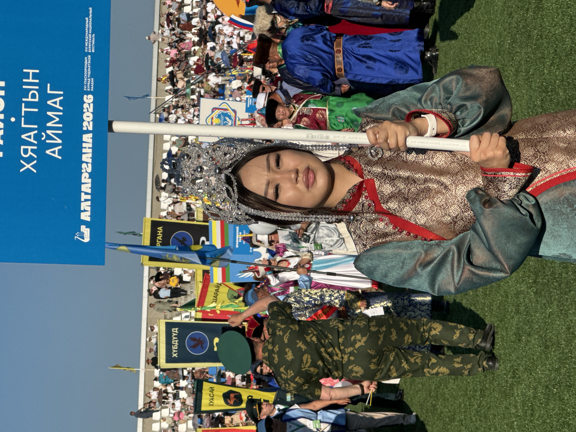 The image depicts a formal procession or event taking place on a sunny day. In the foreground, a young woman adorned in traditional Buryat attire, including an elaborate headdress and embroidered robe, holds a white pole. Her expression is neutral, and she looks directly at the camera. Behind her, a diverse group of people are visible, many wearing traditional clothing or uniforms. Spectators are seated in tiered stadium seating in the background, under a clear blue sky. Flags and banners bearing text in Cyrillic script are displayed, suggesting a cultural festival or national celebration. One prominent blue banner in the upper portion of the image reads "РАЙОН ХЯАГТЫН АЙМАГ" and "АЛТАРГАНА 2026", indicating the location as Kyakhtinsky District and the event as "Altargana 2026". This event is likely the Altargana festival, a major cultural gathering for Buryat people, which often takes place in Buryatia, Russia. The overall scene suggests a vibrant and organized event showcasing cultural heritage.