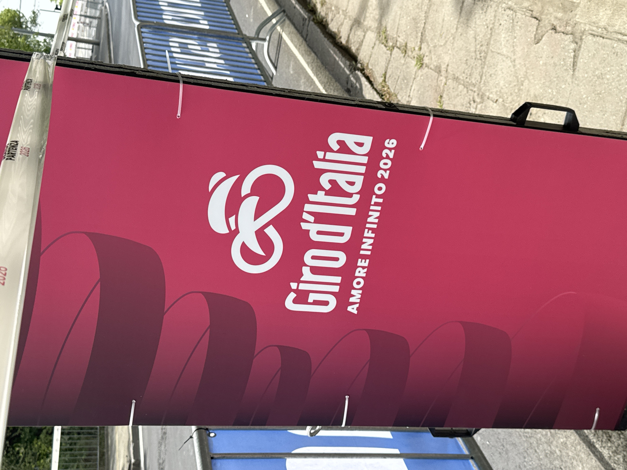 The image shows a sign for the Giro d'Italia Amore Infinito 2026. The sign is predominantly magenta with white lettering and graphics. A series of curved, dark red ribbons runs along the bottom of the sign. Above this is the Giro d'Italia logo, a stylized infinity symbol with the text "Giro d'Italia" stacked to the right. Below this is "AMORE INFINITO 2026." The sign appears to be affixed to a barrier or frame. The background reveals a portion of a pedestrian area, including the city of Burgas, Bulgaria with visible sidewalk and metal railings. The light conditions suggest it is daytime.