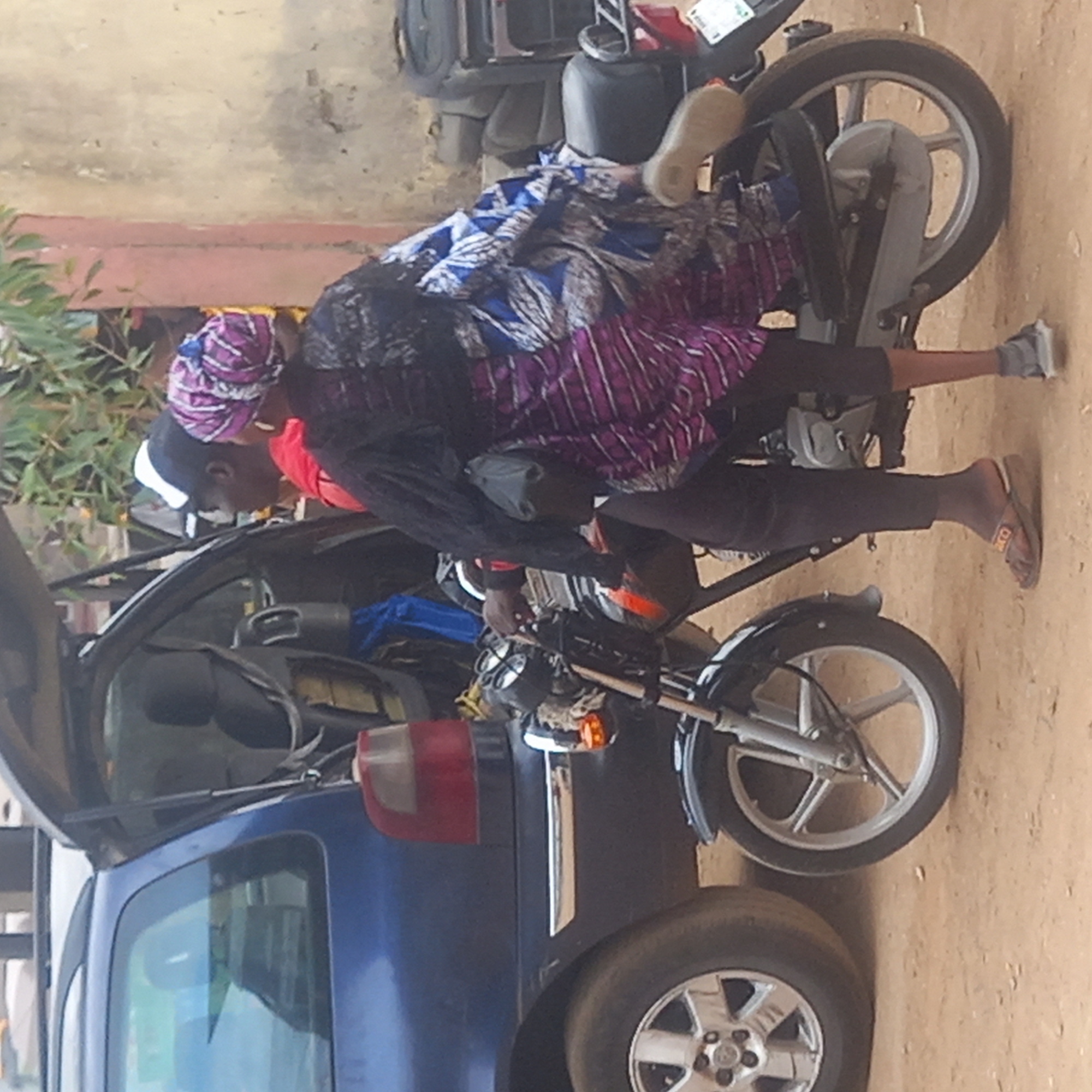 A person in a black long-sleeved shirt and purple striped skirt is riding a motorcycle next to a dark blue car with its hatchback open. The person is wearing a headscarf and sandals. The motorcycle is parked on a dirt surface. The location is Sardauna, Nigeria.