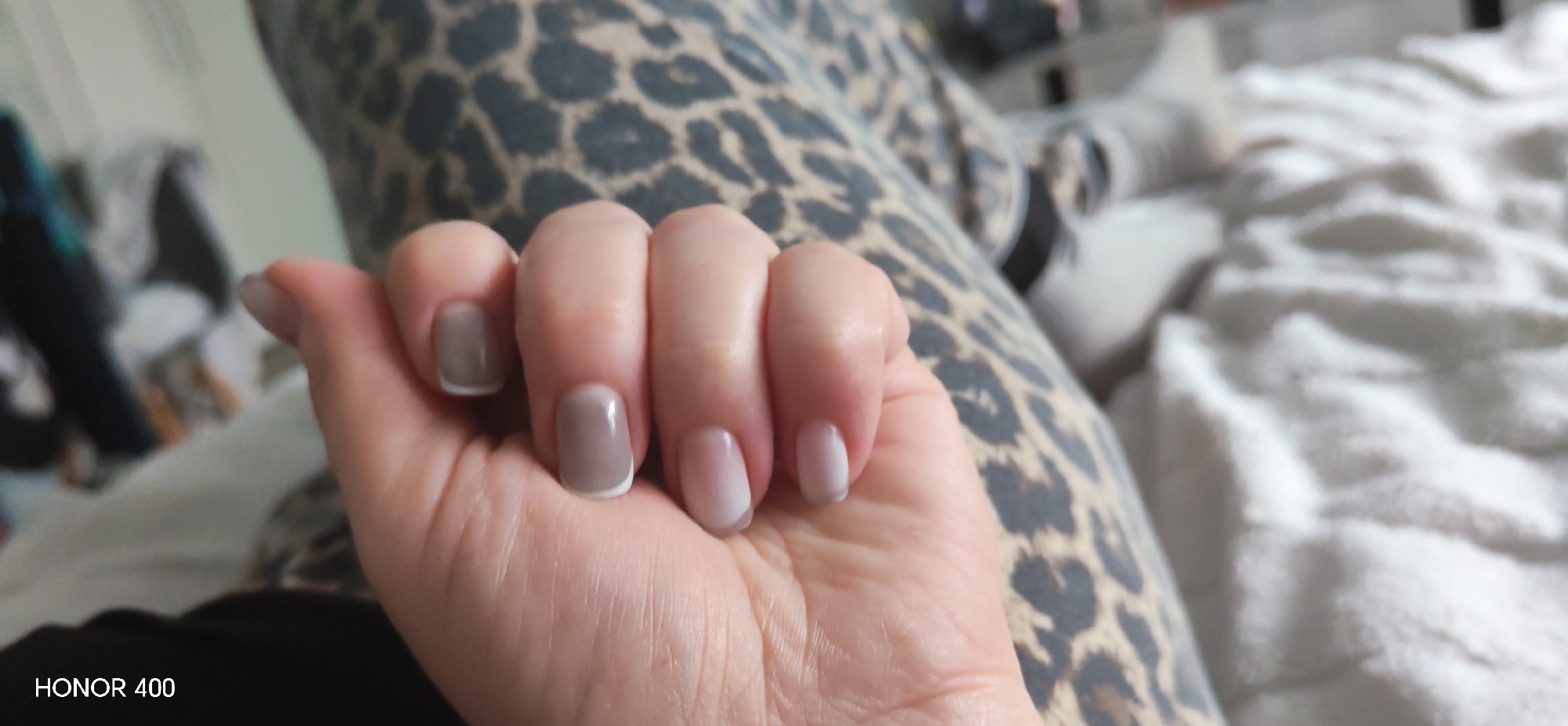 A close-up shot focuses on a person's hand with manicured nails, featuring a gradient effect from a taupe color to a pale white at the tips. The thumb nail has a white edge. The hand rests against a background of leopard-print fabric and a soft white textured blanket. The lower left corner of the image includes "HONOR 400" in white text. The location of Noviy, Russia is not determined within the image, nor do any details suggest it.