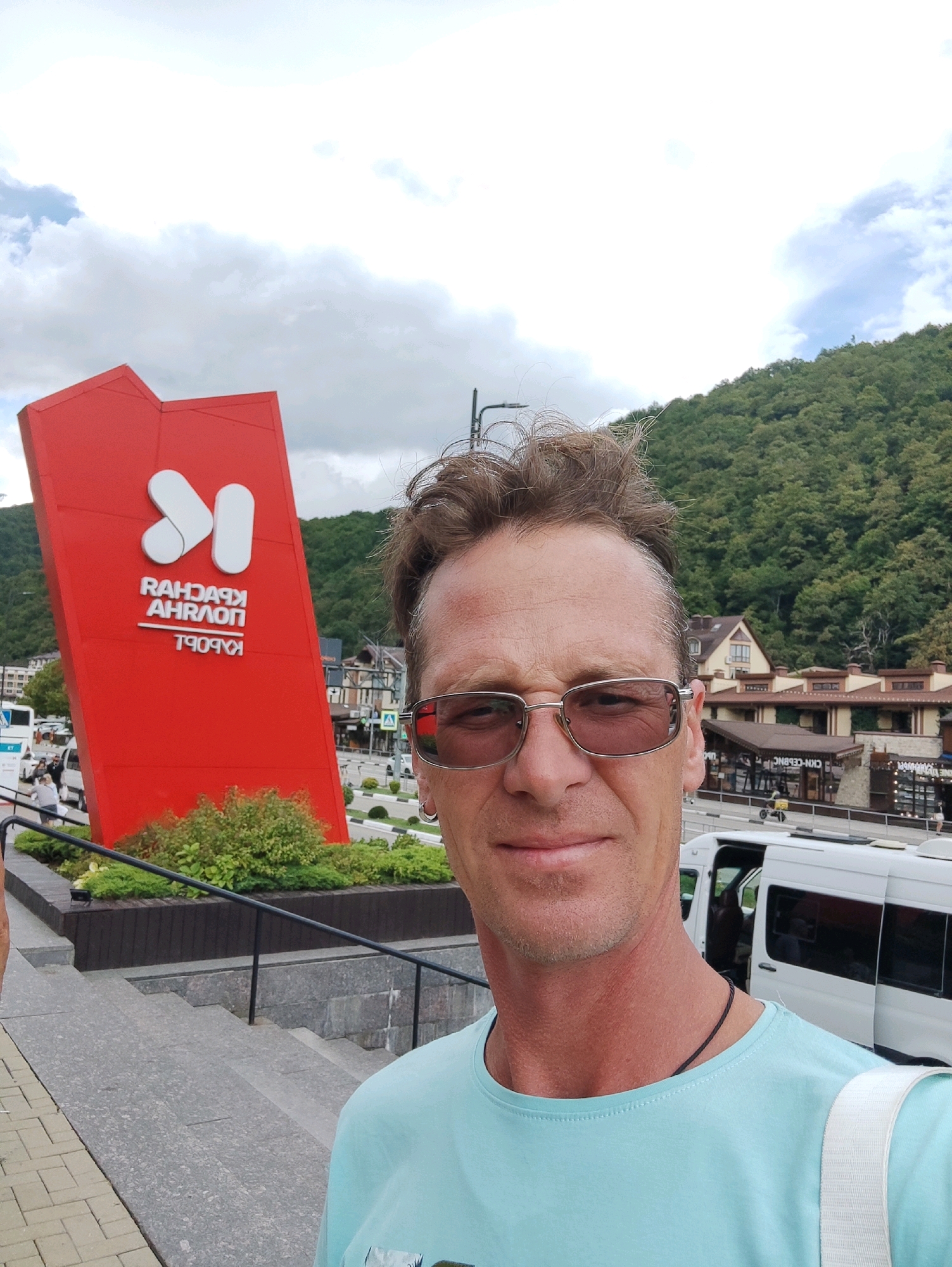 A selfie of a man with short brown hair and sunglasses is taken outdoors in Estosadok, Russia. He is wearing a light blue t-shirt and a dark cord necklace. Behind him, a large red sign with white Cyrillic lettering and stylized white shapes stands prominently. The sign reads "КРАСНАЯ АХРАЛON" and "ТРОЯН". In the background, green trees cover a mountainside, and several buildings with brown roofs are visible. White vans are parked on a street to the right of the sign. A set of stone steps leads down to the left of the frame.