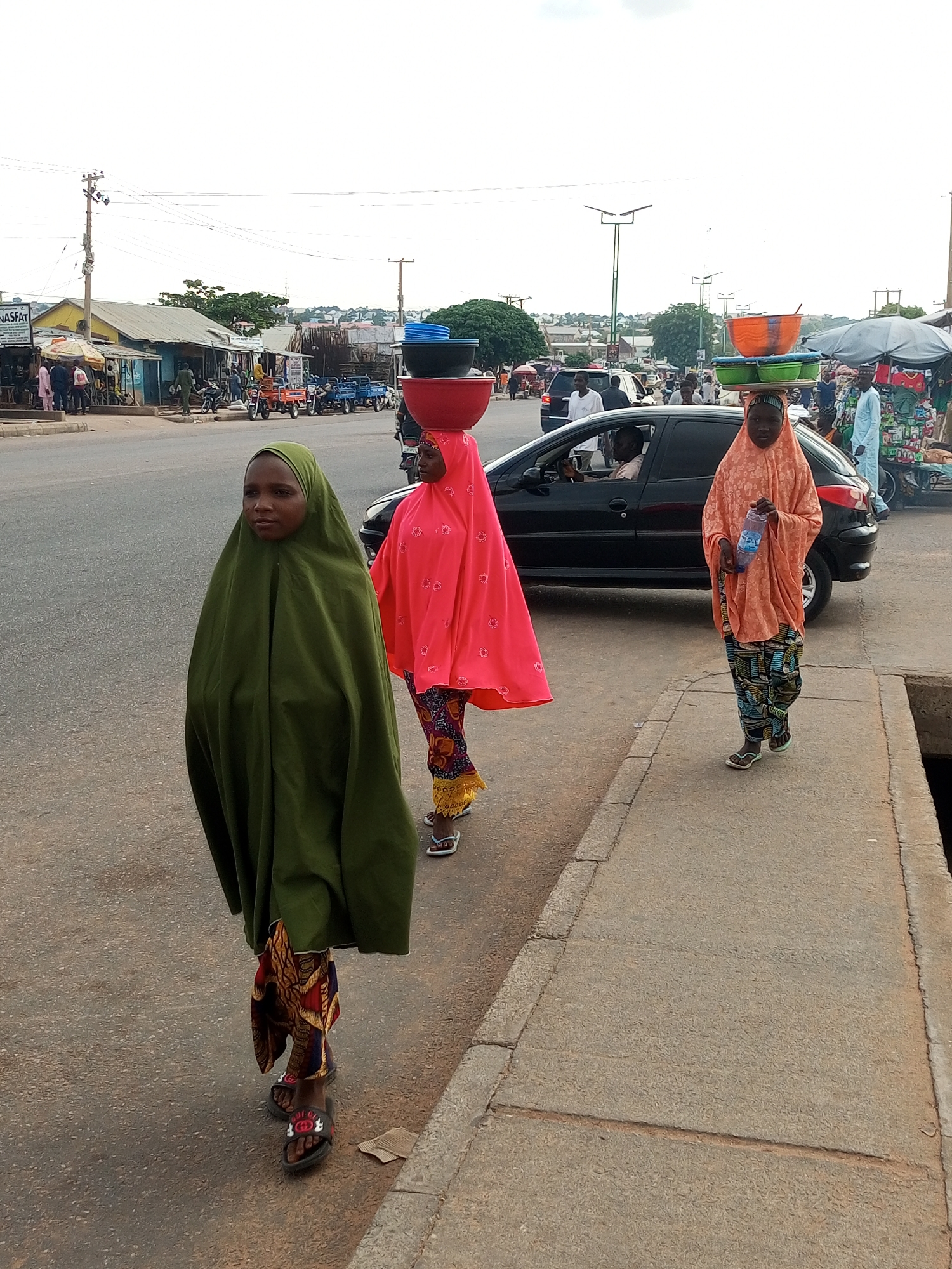 In an outdoor urban environment, possibly a marketplace, three young females walk along a paved sidewalk adjacent to a street. The female in the foreground, on the left, is clad in a long, dark green hijab and a patterned cloth skirt. She wears black sandals with red accents. Next to her, to the right, a female in a vibrant pink hijab and a patterned skirt walks forward, also wearing sandals. She appears to be carrying a stack of colorful bowls on her head, with the uppermost bowl being red. Further to the right, another female in an orange hijab and a patterned skirt holds a plastic water bottle. She also appears to be carrying a stack of bowls on her head, with the top being orange, followed by green and a dark blue bowl. The street next to the sidewalk features several vehicles, including a black car with a driver visible inside, and various parked motorbikes and carts in the background. Buildings and market stalls line the opposite side of the street. The sky is overcast.