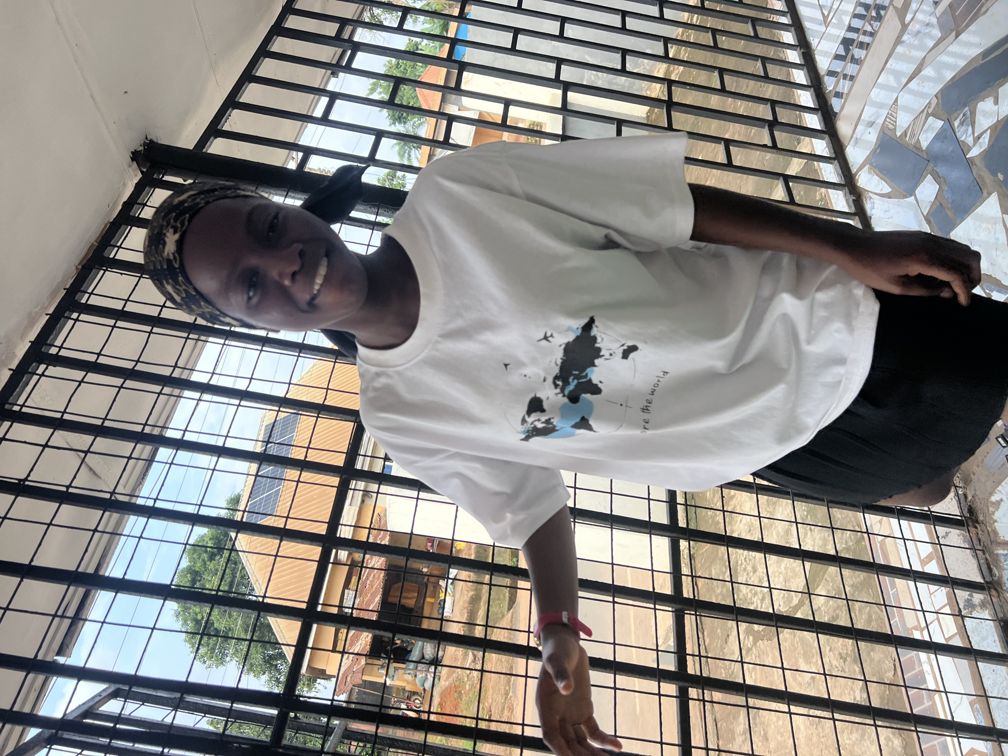A young Black woman with dark skin and a bandana covering her hair is standing outside, leaning against a black metal fence. She is wearing a white t-shirt with a graphic of the world map on it and a black skirt. She is smiling and looking at the camera. Behind her, through the fence, is a building with a brown roof and solar panels, a large tree, and a cloudy sky. The ground in front of her is tiled with a mosaic pattern of blue, gray, and white squares. The scene appears to be in Nkpor, Nigeria, given the architecture and vegetation.