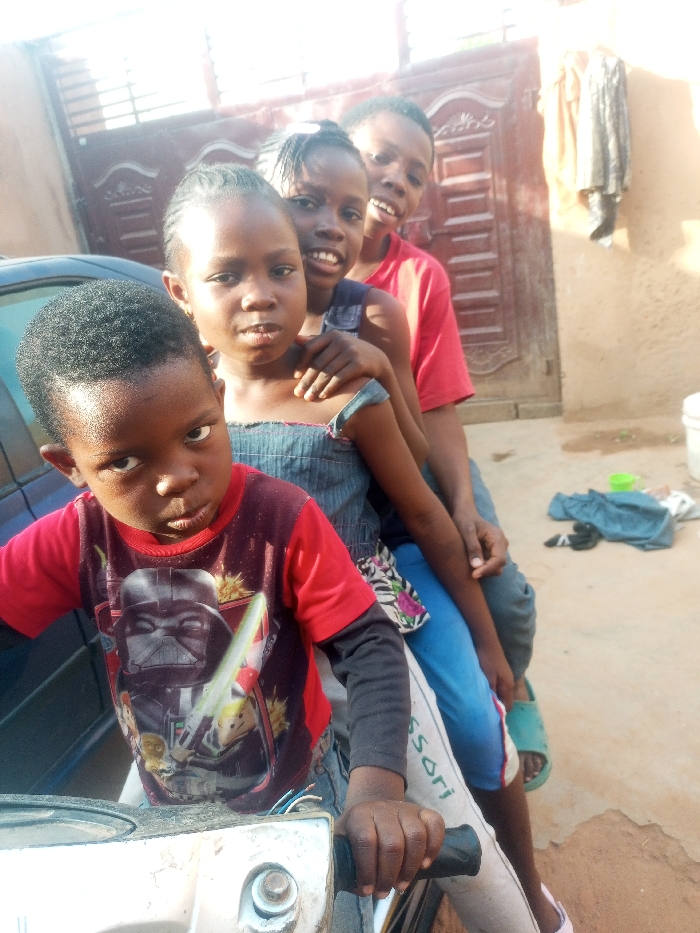 Four children are lined up on a motorcycle. The child in front wears a red shirt with a Star Wars graphic. Behind him is a girl in a denim top. Further back is another girl smiling with an arm around the first girl. The last child, a boy in a red shirt, smiles slightly. The background features a closed gate and a wall with hanging clothes. Casual attire and bright daylight suggest a relaxed, outdoor setting.