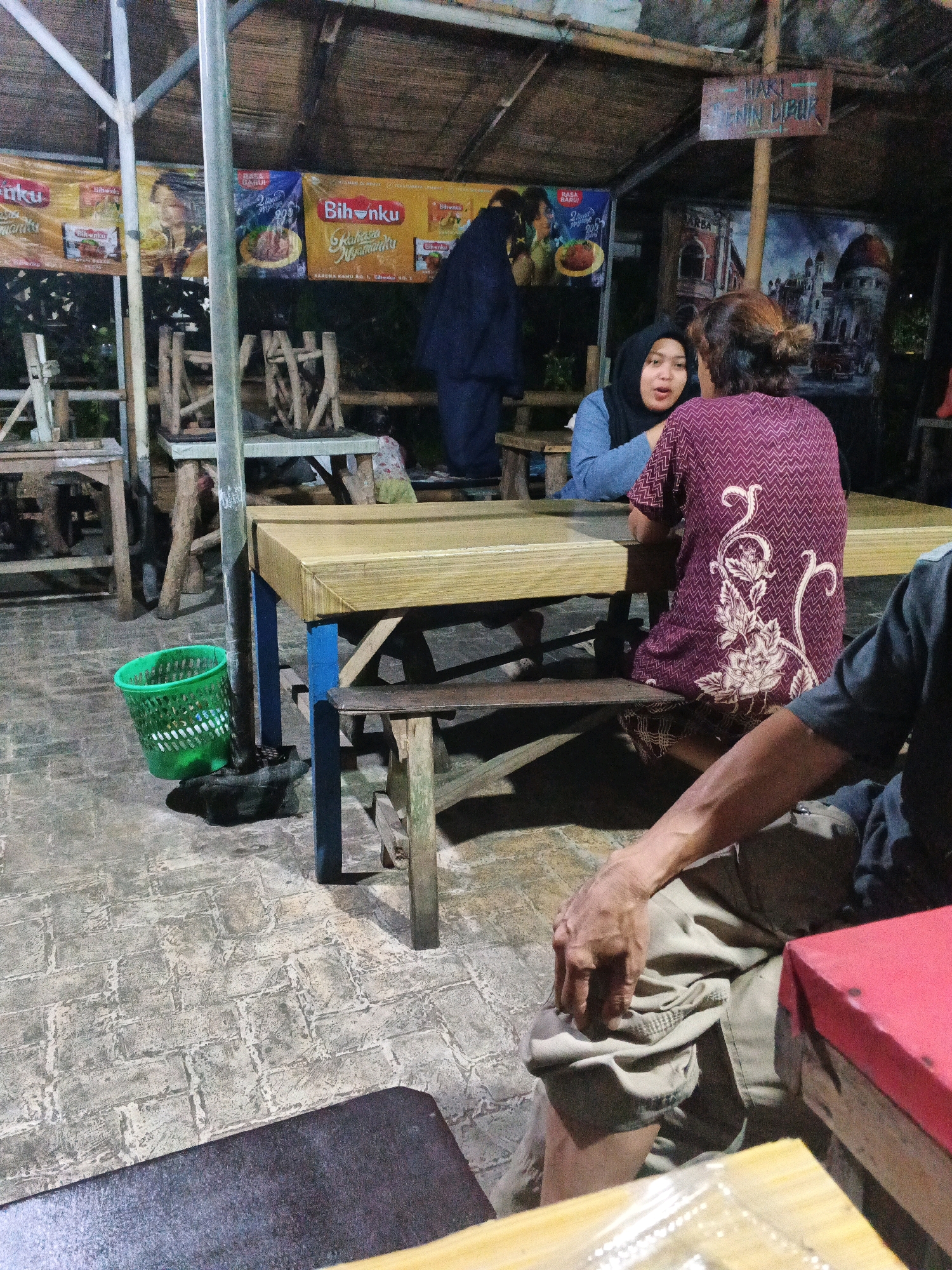 The image depicts an outdoor seating area of a food establishment, possibly a warung or a small restaurant, located in Ungaran, Indonesia. The scene is illuminated by artificial lighting, suggesting it is evening or night. In the foreground, a person's arm and legs are visible, wearing khaki pants and resting on a table. They appear to be sitting at a table with a red box on it.
Behind this person, a wooden table with a blue leg structure is prominent. Two women are seated at this table, facing each other and appearing to be in conversation. One woman is wearing a dark blue hijab and a light blue top, while the other is wearing a patterned purple top.
The background reveals a simple roof structure made of woven material supported by metal poles. Several banners advertising food products are displayed on the wall behind the tables. Wooden chairs and tables, some appearing rustic and made of branches, are scattered throughout the area. A green plastic trash can is placed near a pole supporting the roof. The ground is paved with what appears to be stone or concrete tiles arranged in a brick pattern. In the distance, murals or paintings adorn the walls, one depicting a cityscape and another a natural scene. A sign on the wall reads "HARI SENIN LIBUR" (Monday is holiday).