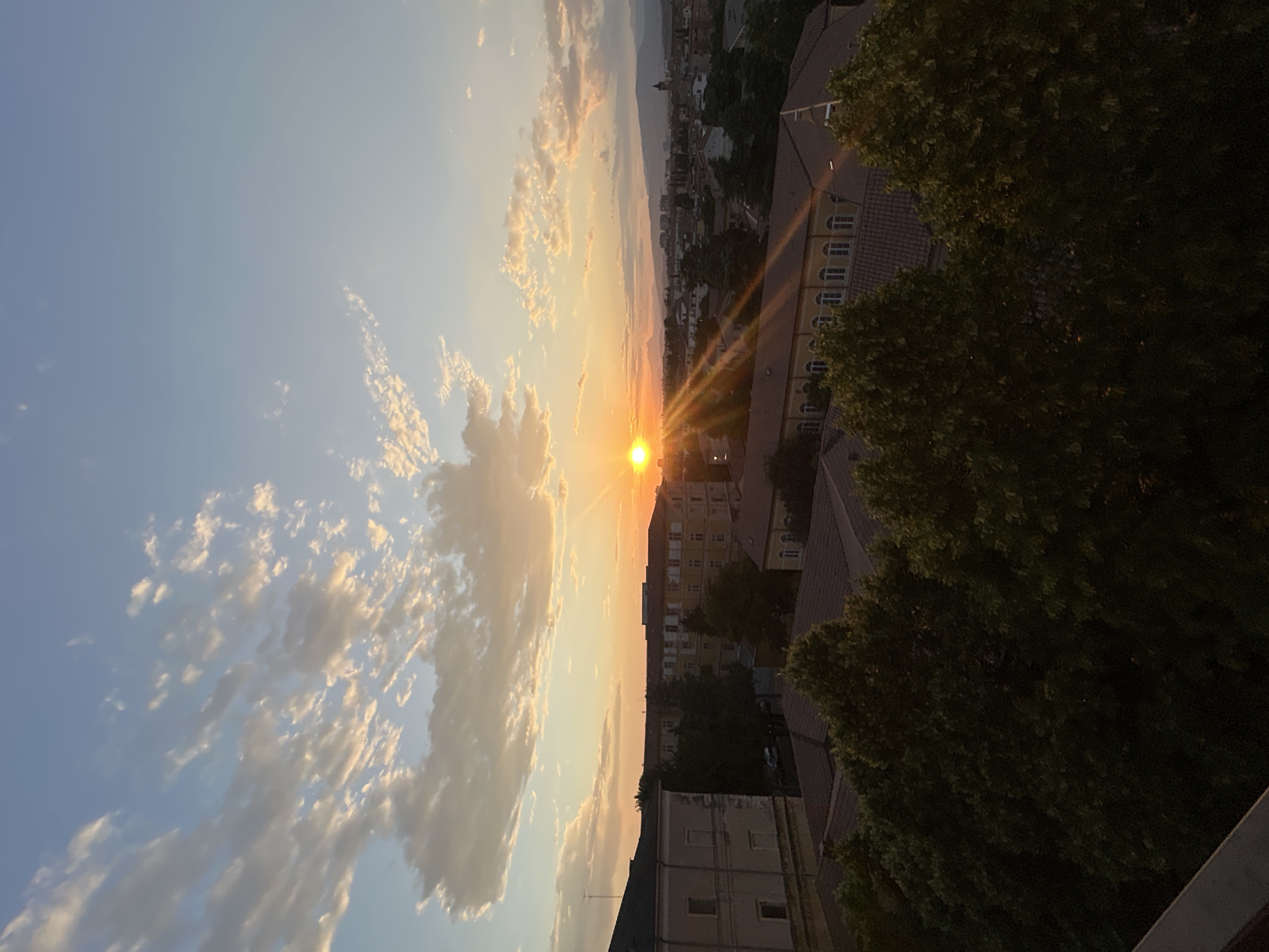 The image shows a sunset over the city of Verona, Italy. The sun is low on the horizon, casting a warm orange glow over the sky and the buildings below. In the foreground, there are trees, and in the middle ground, there are several buildings with red tile roofs. In the background, there are more buildings, and mountains can be seen in the distance. The sky is filled with scattered clouds that are illuminated by the setting sun. The overall scene is peaceful and serene. The image appears to have been taken from a high vantage point, possibly a balcony or a rooftop.