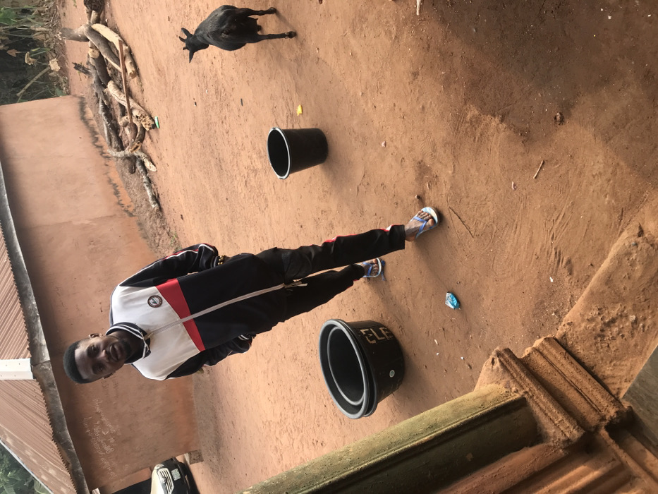 A young adult male is centrally positioned outdoors in Ogodo, Nigeria, standing on reddish-brown dirt. He wears a color-block track jacket (white, red, black), black track pants with red stripes, and light blue and white flip-flops. His hands are in his pockets, and he smiles slightly while looking directly at the camera. A gold-colored bracelet is visible on his right wrist. To his left, two black plastic buckets are nested, with the word "ELF" visible on the upper bucket. A single black plastic bucket is positioned to his right. A black goat with visible horns stands towards the right, facing away from the camera. In the background, a one-story building with a light brown wall and a corrugated metal roof is visible, with a pile of cut tree branches and logs to its right. A silver vehicle is partially visible on the far left. Scattered on the dirt ground are a crumpled light blue wrapper, a small yellow piece of debris, and other small natural fragments. A textured, reddish-brown architectural element occupies the lower left foreground.