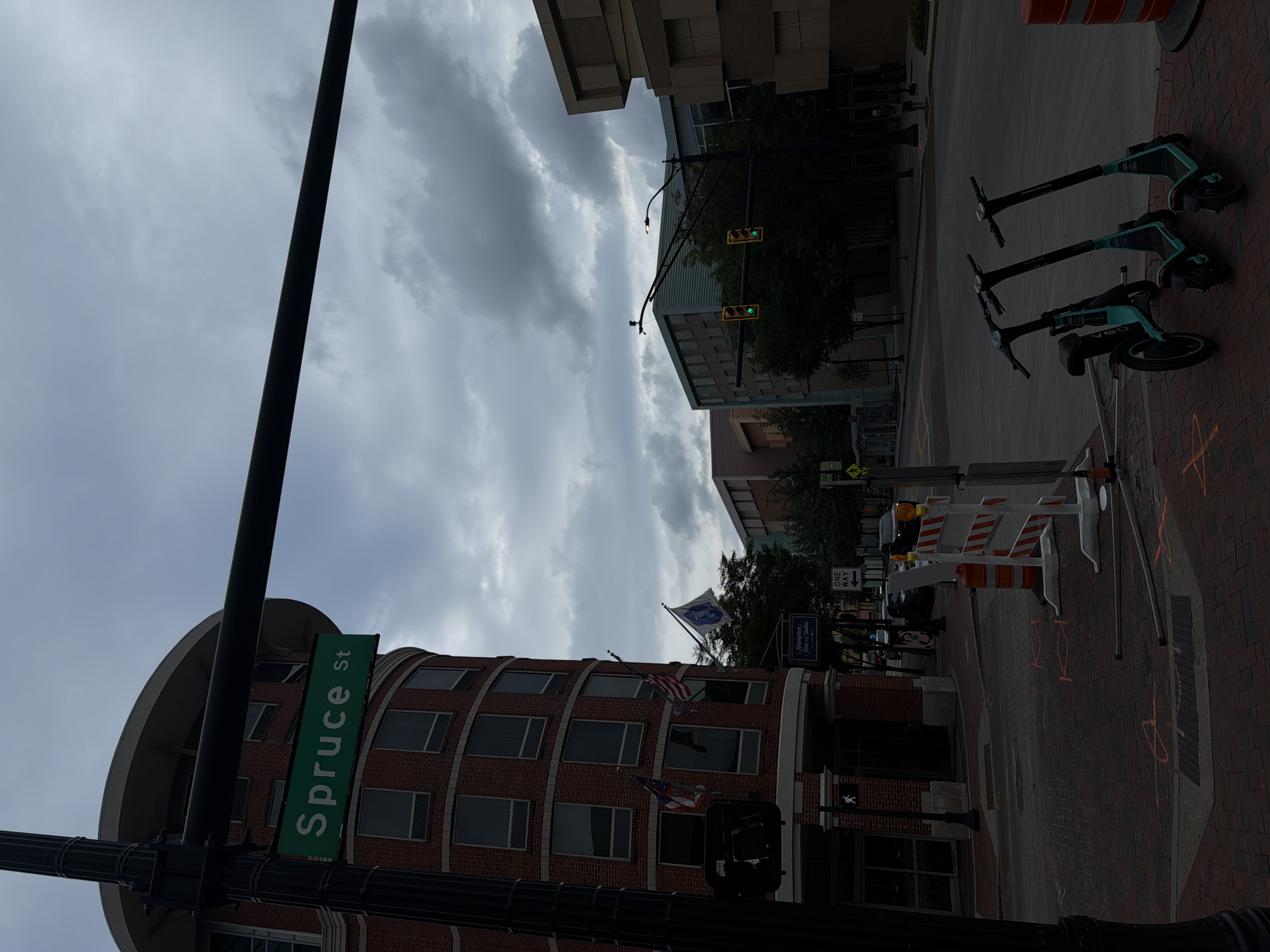This is an image of an overcast day in Columbus, Ohio. The main subject is a street sign that reads "Spruce St" on a green background. To the left of the sign is a brick building, likely a hotel, as suggested by a sign for "Hampton Inn & Suites." In front of the hotel, there is a pedestrian crossing signal and a traffic light. On the sidewalk in front of the building, there are three electric scooters lined up. The street is paved with brick and there are orange markings on the ground, indicating construction or utility work. In the background, there are several modern buildings and trees. A "One Way" sign is visible pointing to the right.