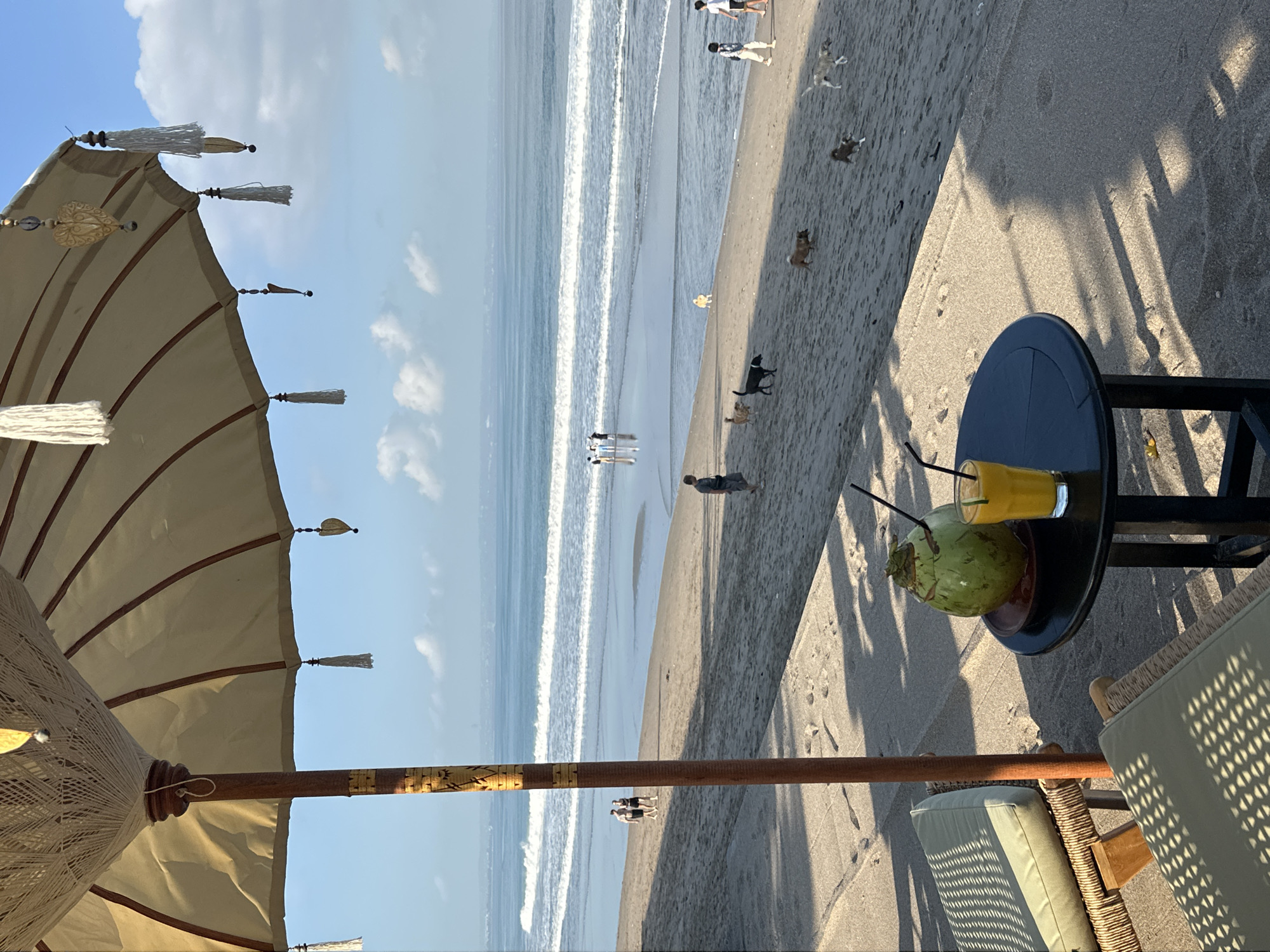 A beachside scene in North Kuta, Indonesia, features a large, decorative umbrella casting shadows on two lounge chairs. In the foreground, a small black table holds a fresh coconut and a glass of orange juice with straws. Beyond the chairs, the sandy beach is populated with several people and dogs walking and playing near the gentle ocean waves. The sea stretches to the horizon under a partially cloudy sky.
