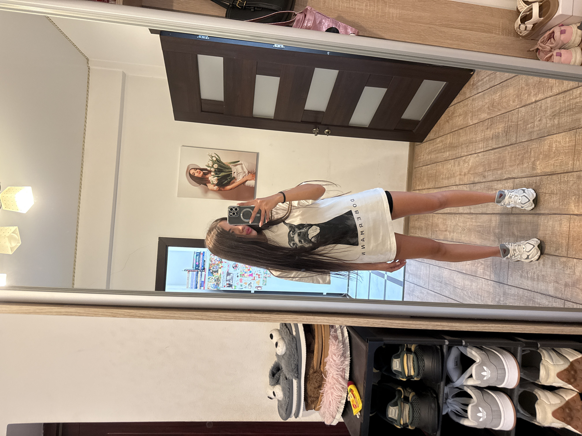 A young woman with long dark hair is taking a mirror selfie in a room. She is wearing a white t-shirt with a graphic print of a Doberman Pinscher and black shorts. She is standing in front of a dark brown wooden door with frosted glass panels. To her left, there is a tall mirror with a light wood frame. Next to the mirror is a shoe rack with several pairs of shoes and slippers. On the wall behind her, there is a framed picture of a woman holding a bouquet of tulips. The room has wooden flooring and a white ceiling with lighting fixtures. The woman's posture and the reflection in the mirror suggest she is indoors, likely in a residential setting. The specific location within Chișinău, Moldova cannot be determined from the provided image.
