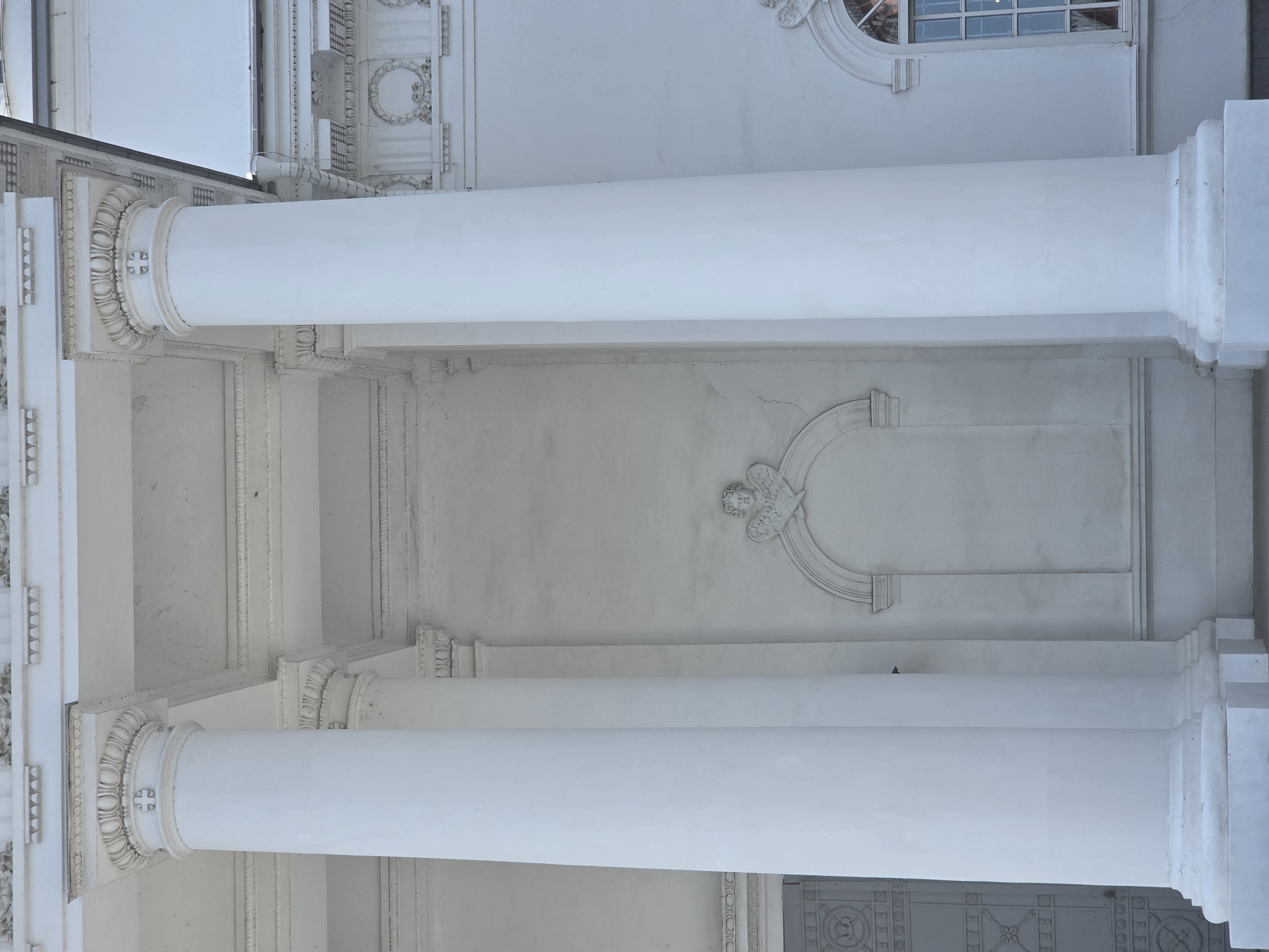 The image shows a close-up, low-angle view of the exterior of the Main Cathedral of Alexander Nevsky in Nizhny Novgorod, Russia. The scene is dominated by two massive white Doric columns in the foreground, their fluted shafts and ornate capitals rendered in sharp detail. Between these columns, a section of the white facade features an arched niche with decorative relief work above it, depicting a cherub's head with wings. The architectural style is classical, with detailed molding and friezes visible along the upper parts of the building. A segment of a window with multiple panes is visible on the right side, its glass reflecting the sky and surrounding structures. The overall impression is one of grandeur and solemnity, with the bright white of the building contrasting with the clear sky, suggesting a sunny day. The image is devoid of any human presence or activity, focusing entirely on the architectural elements of the cathedral.